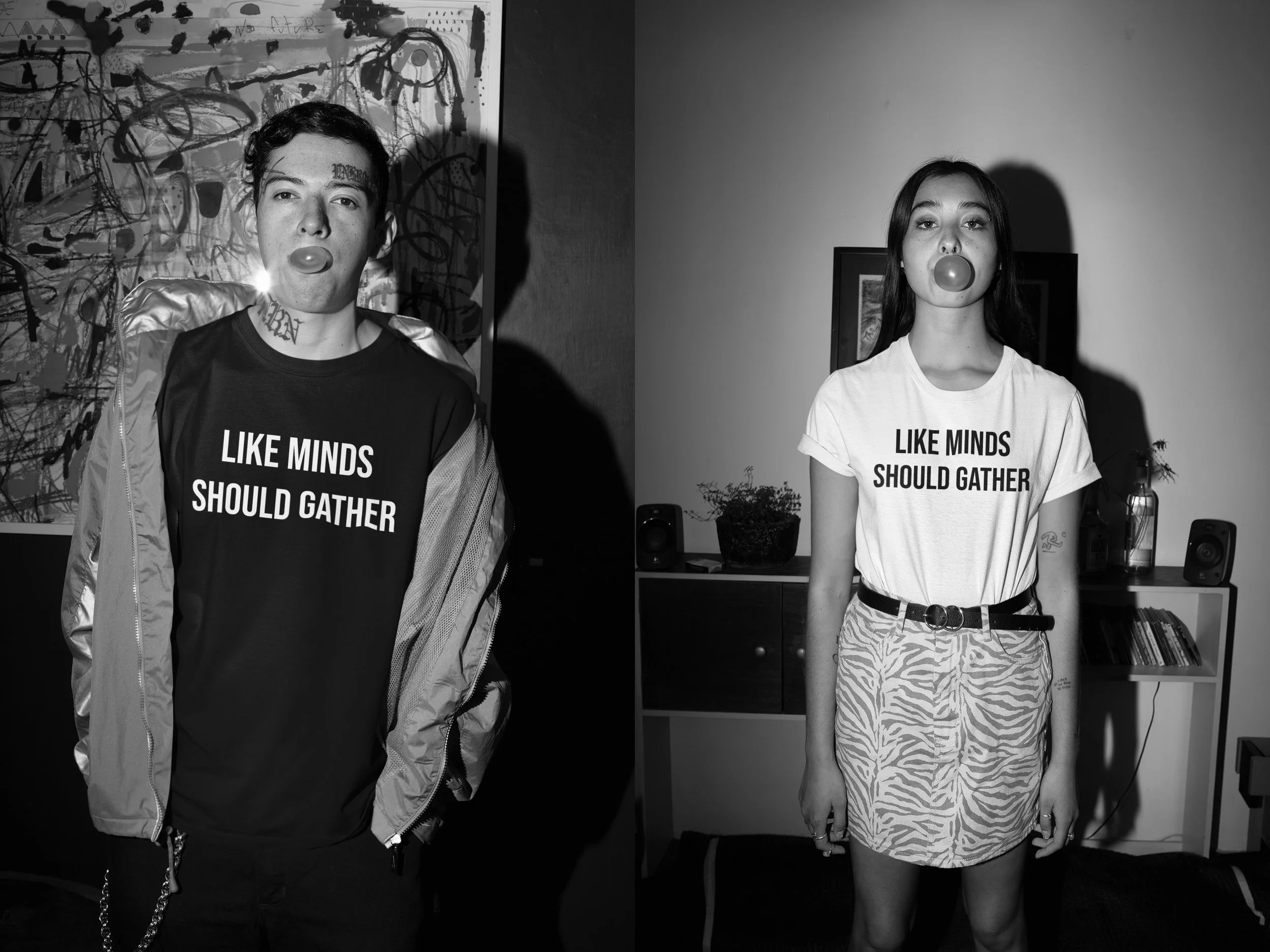 Two young women standing indoors side by side, both blowing bubble gum bubbles and wearing T-shirts that say 'LIKE MINDS SHOULD GATHER' in bold letters. The woman on the left has short hair, tattoos, and is wearing a jacket over her T-shirt, while th