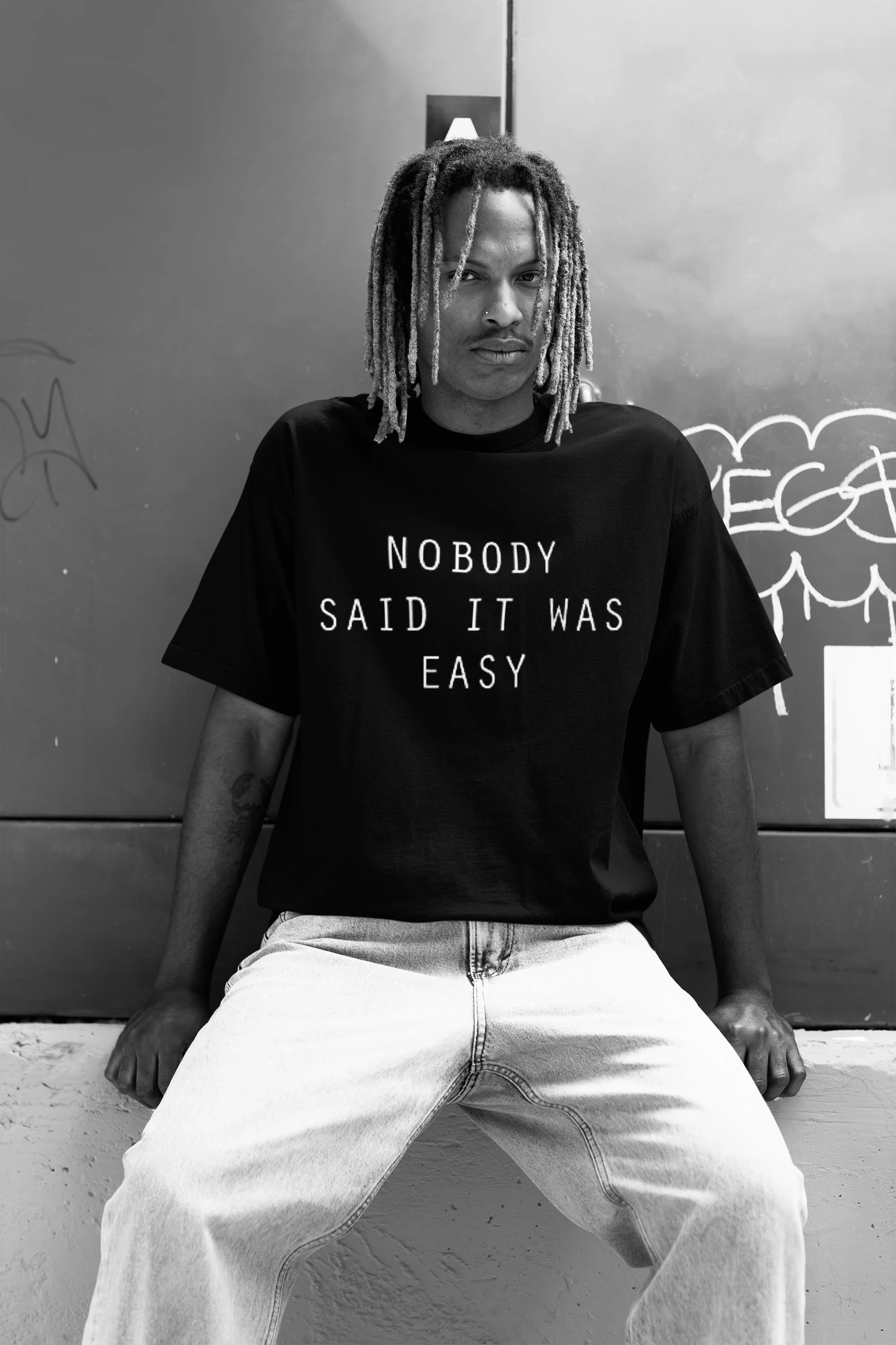 A person with dreadlocks sitting against a wall, wearing a black t-shirt that says 'Nobody Said It Was Easy' in white letters.
