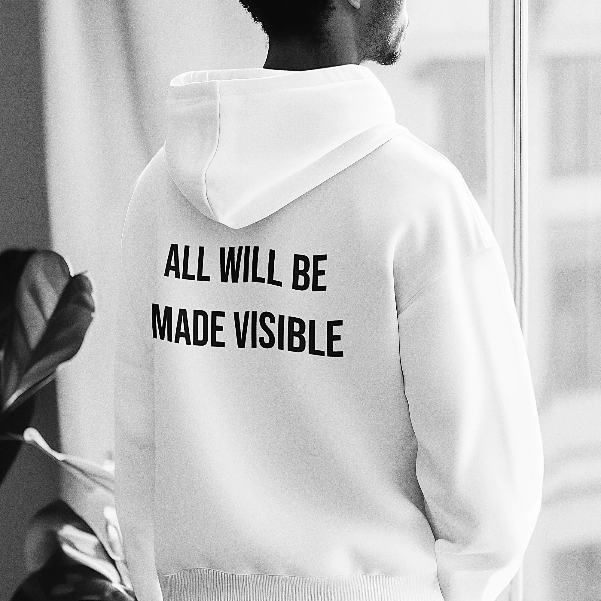 A person wearing a white hoodie with bold black text on the back that reads "ALL WILL BE MADE VISIBLE."