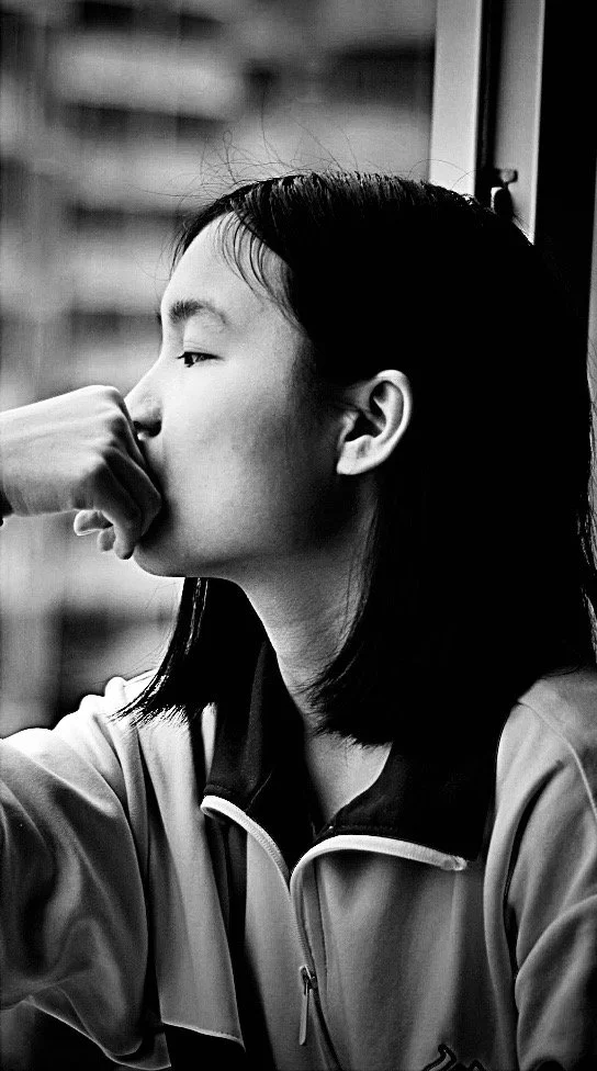 A young woman with shoulder-length hair sitting near a window, resting her chin on her hand, looking outside thoughtfully, in black and white.