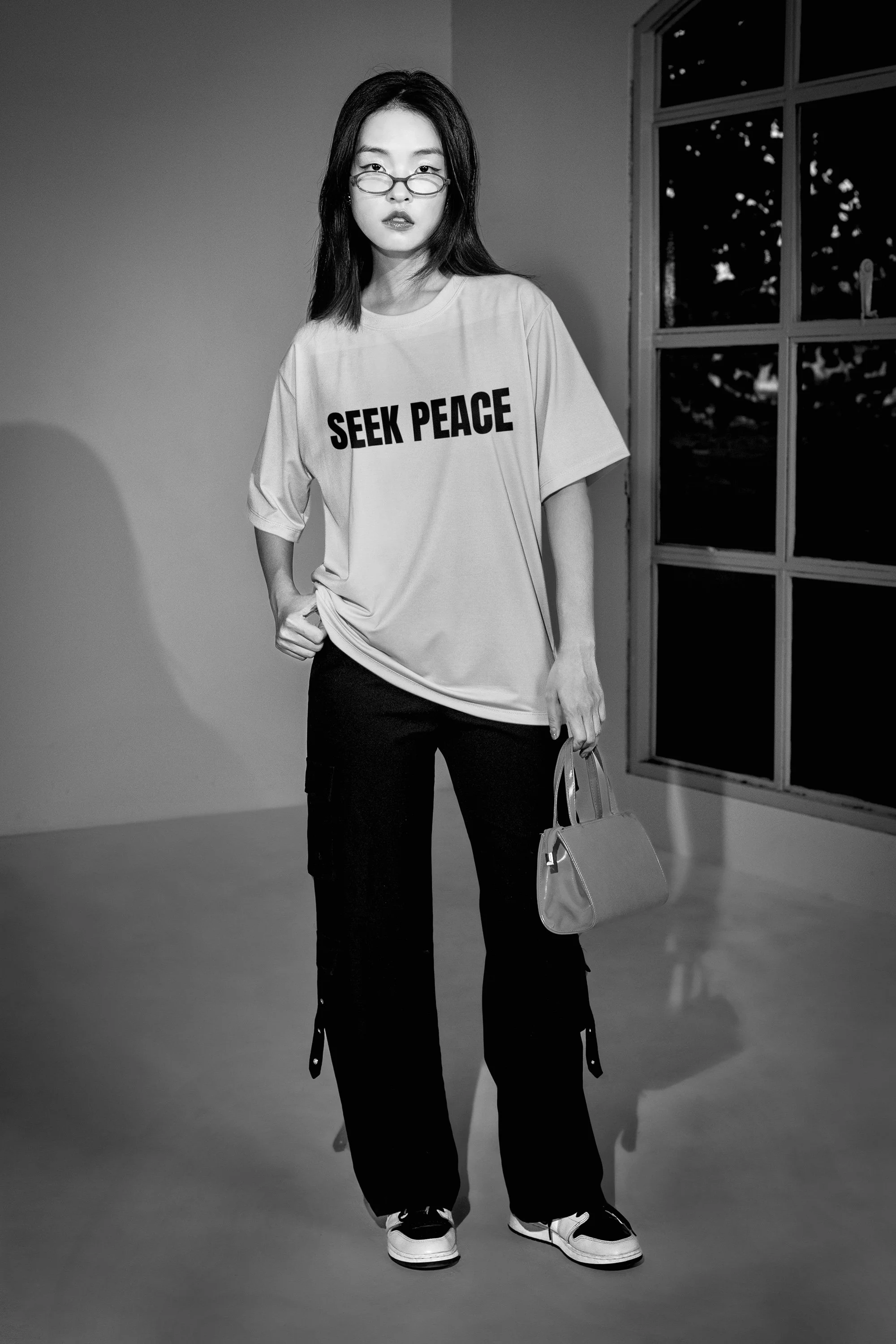 Black and white photo of a woman wearing glasses, an oversized t-shirt with 'SEEK PEACE' written on it, black pants, and sneakers, standing indoors near a large window.
