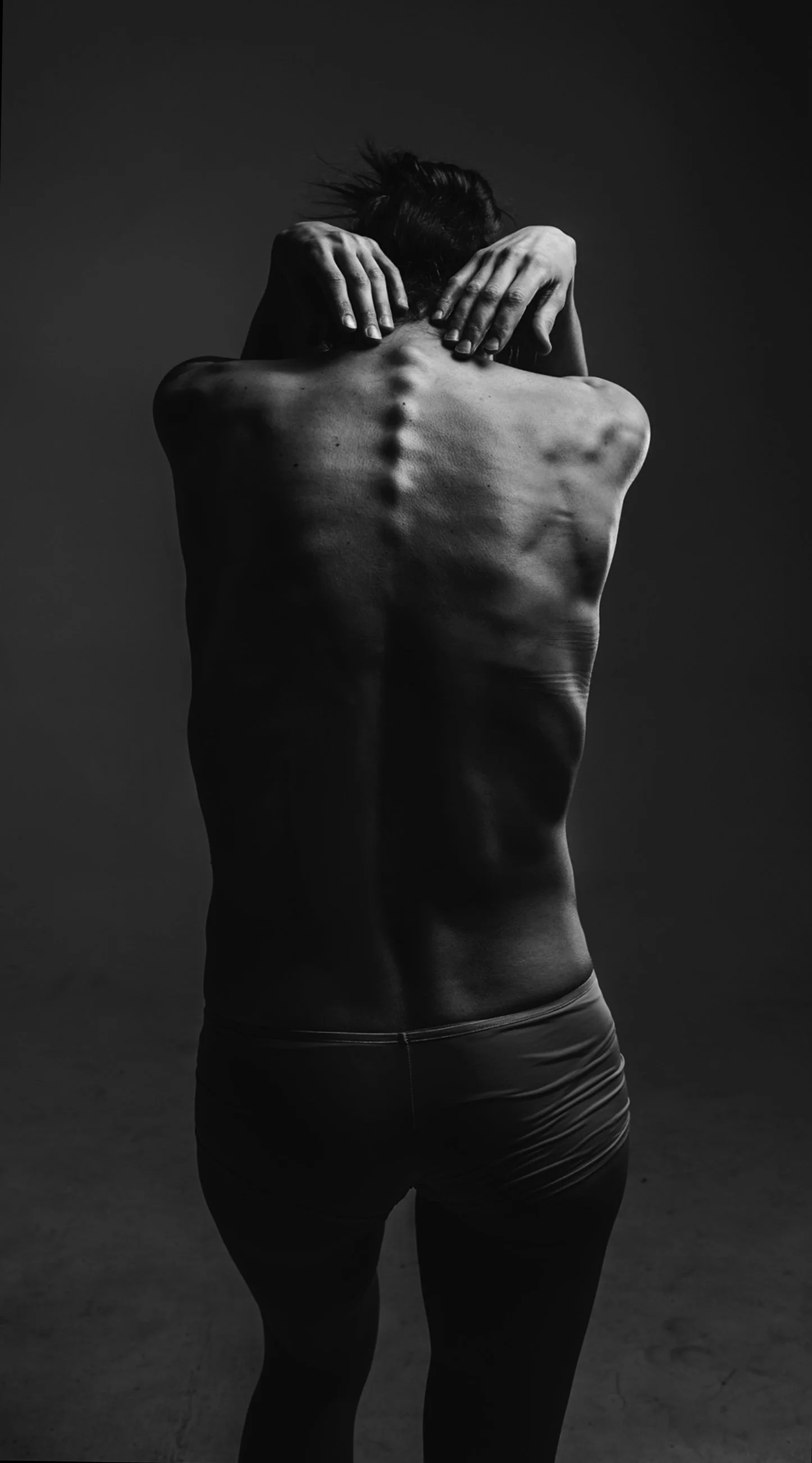 Black and white photo of a woman with her back to the camera, her hands behind her head, showing her spine and shoulder muscles.