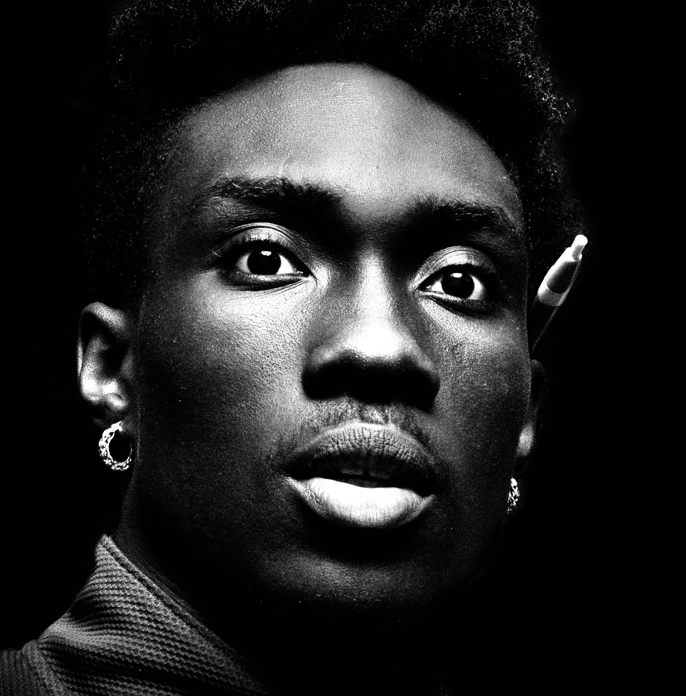 Close-up black and white portrait of a woman with short natural hair wearing earrings, with an eye pin accessory on her right side of the head, against a dark background.