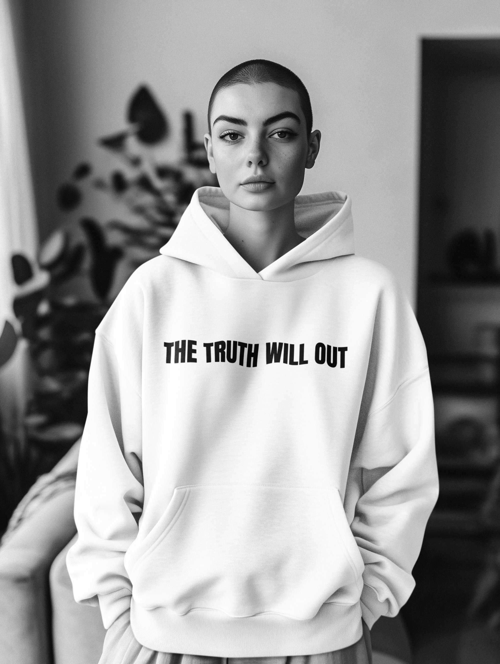 A young woman with a shaved head wearing a white hoodie that says "THE TRUTH WILL OUT" in black letters.