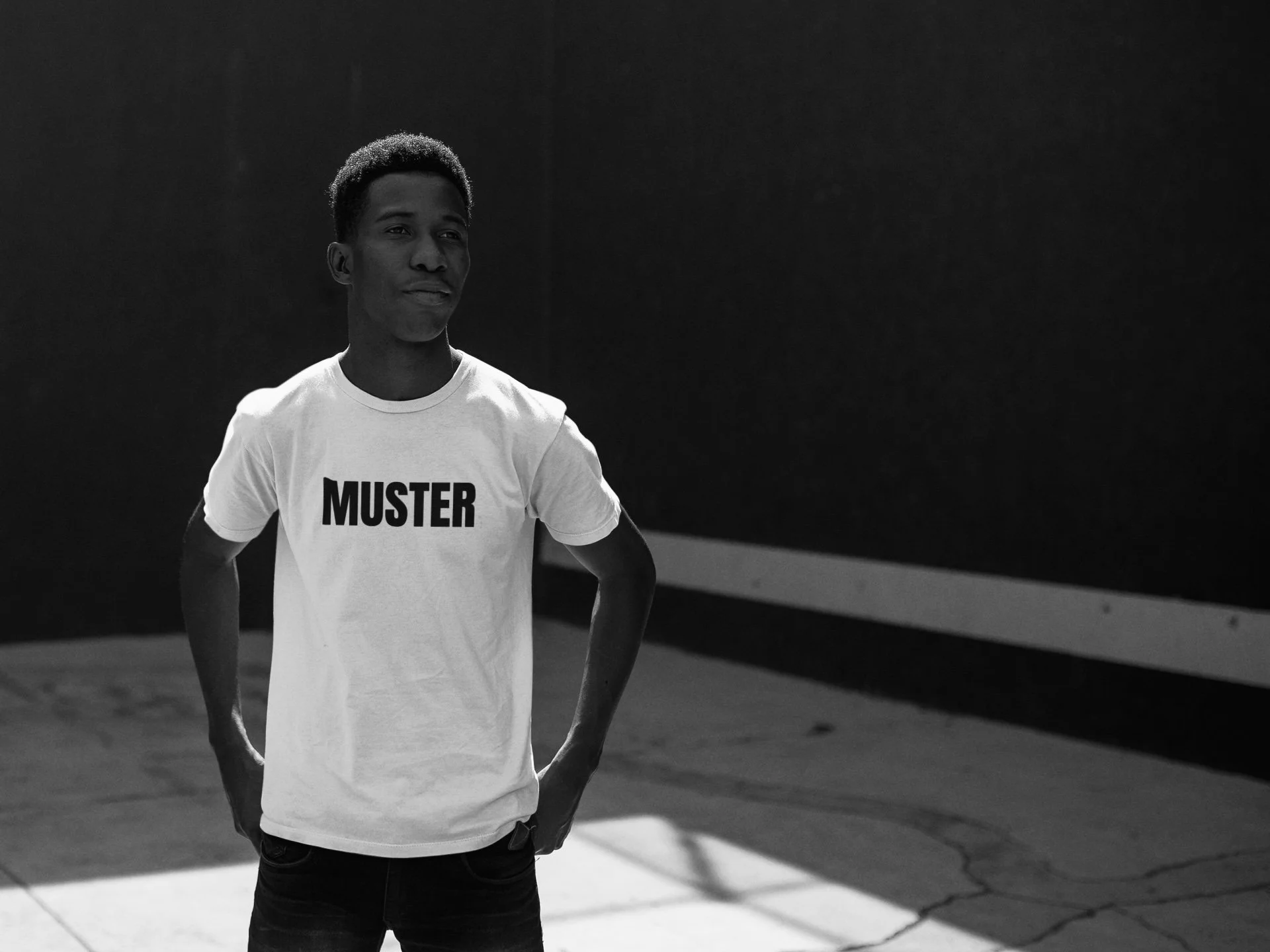 Black and white photo of a young man standing outdoors with hands in his pockets, wearing a white T-shirt with the word 'MUSTER' printed on it.