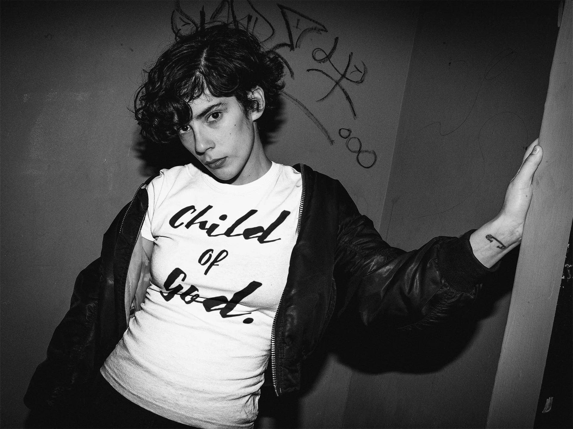 A woman with wavy hair wearing a white t-shirt with the text "child of God" and a black jacket, standing against a wall with graffiti, with her hand on the wall and looking at the camera.