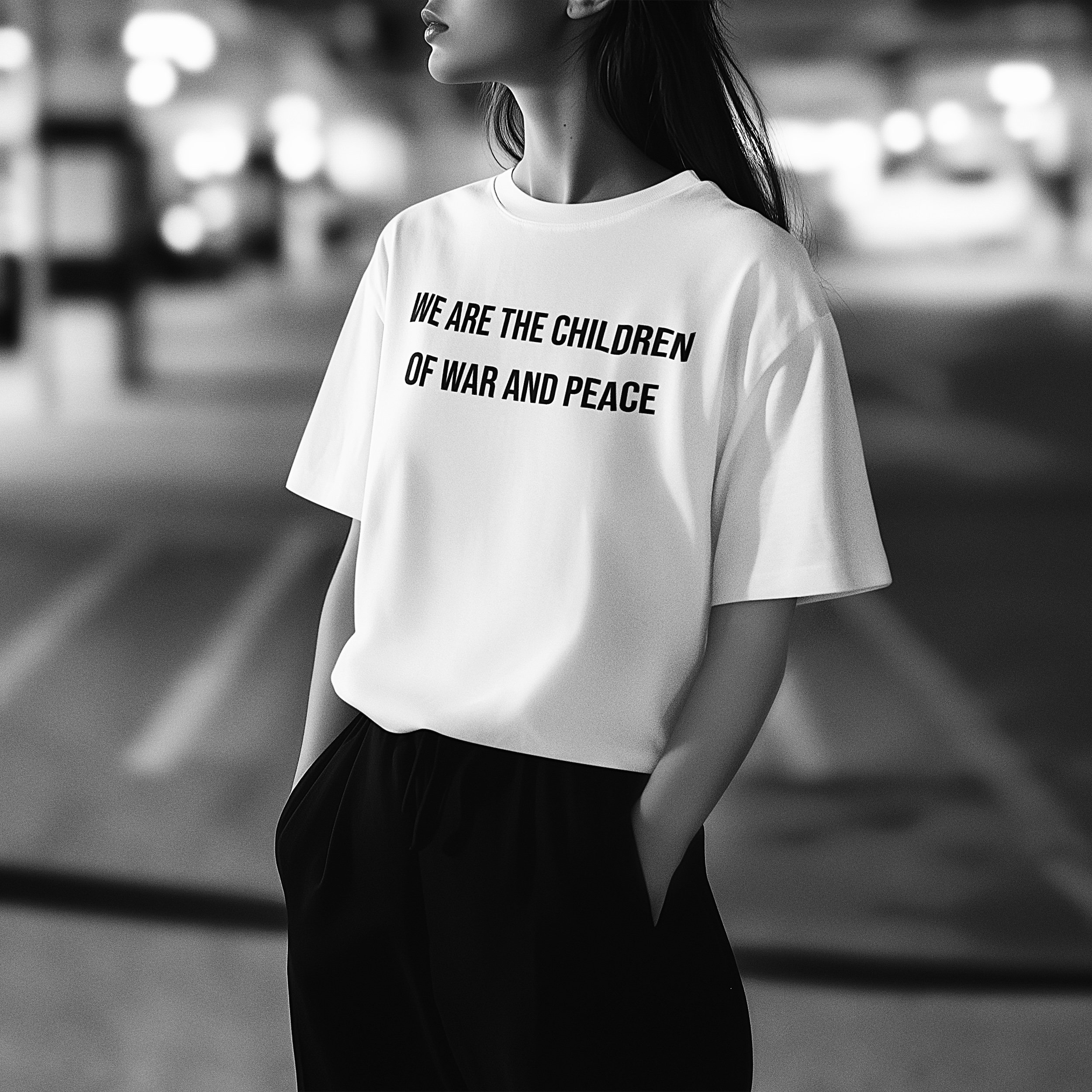 A person standing in an urban setting wearing a white T-shirt with the message "We are the children of war and peace" printed on it, captured in black and white.
