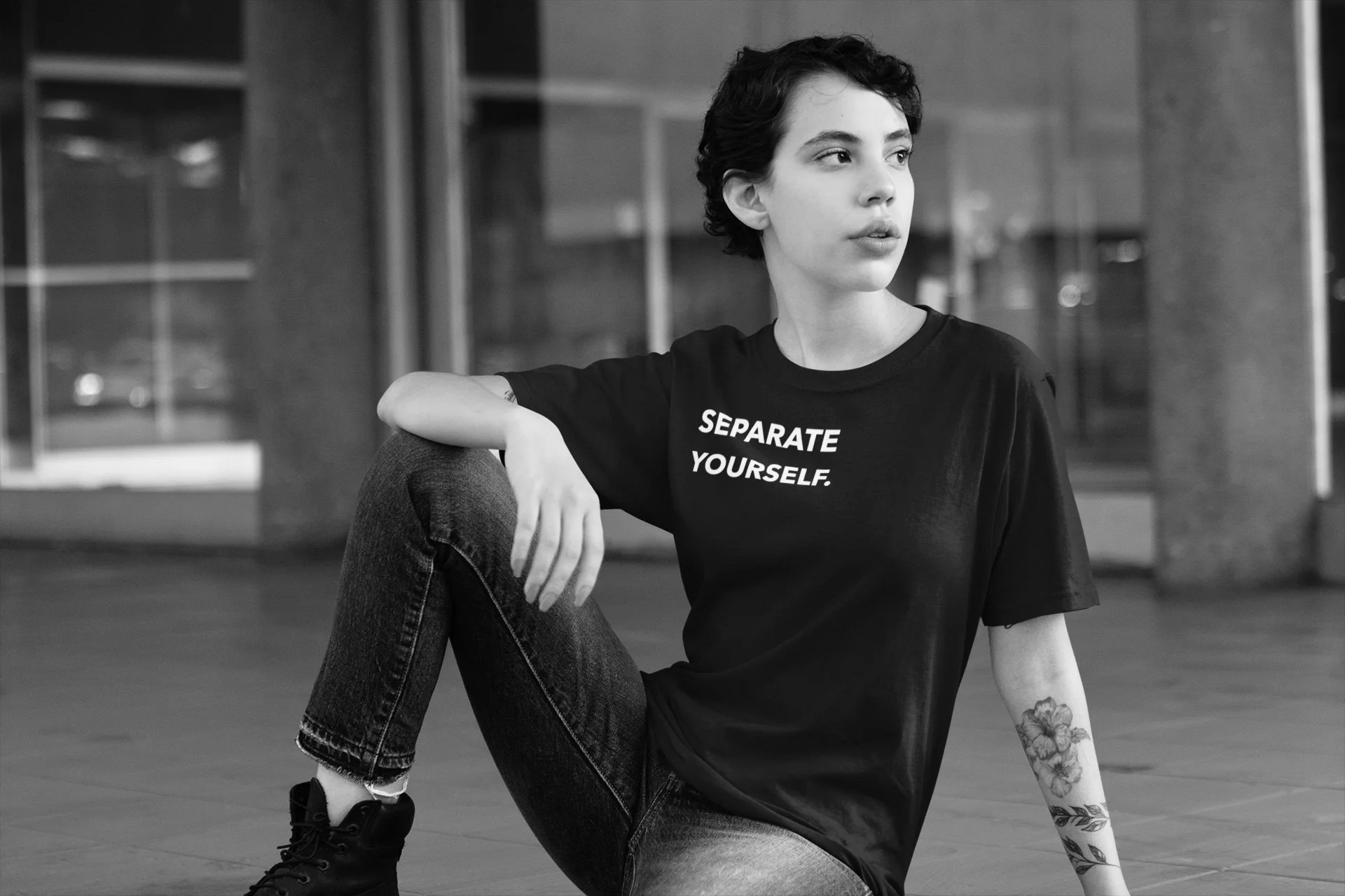 A woman with short curly hair sitting on the floor indoors, wearing a black t-shirt that says 'SEPARATE YOURSELF,' and jeans, with tattoos on her left arm, looking to her right.
