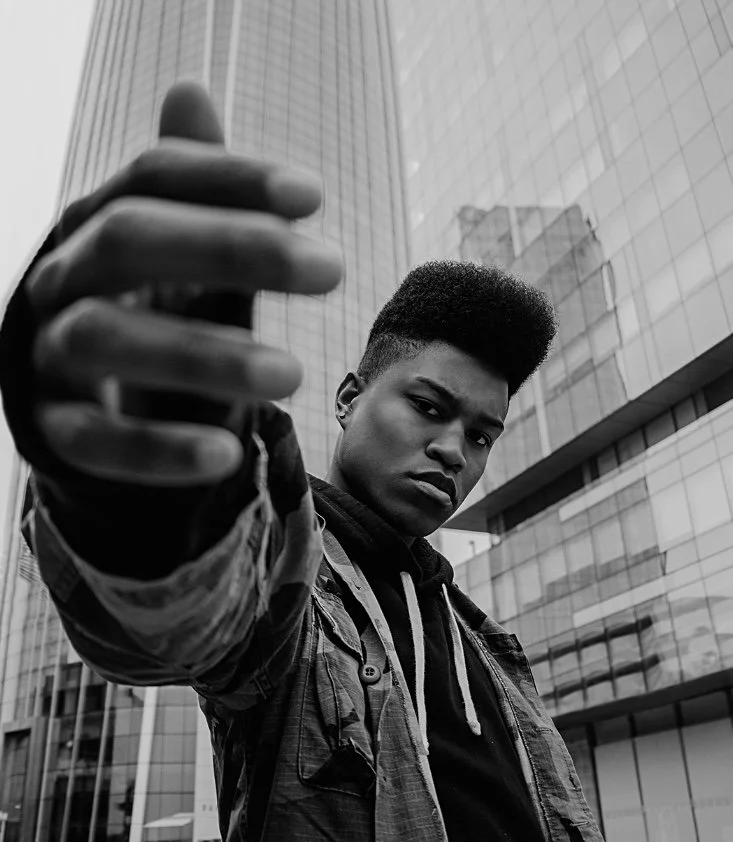 A young man with a flat-top haircut pointing directly at the camera with a serious expression, standing in front of modern glass buildings.