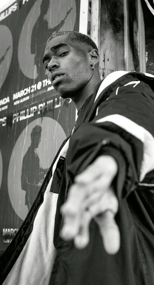 Black and white photo of a young man with short hair, hoop earrings, and wearing a jacket, pointing towards the camera, standing in front of a poster with silhouettes and event details.