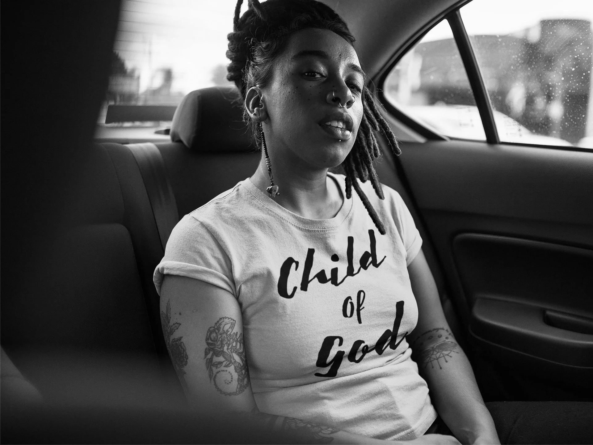 A woman with dreadlocks and tattoos, wearing a T-shirt that says 'Child of God,' sitting in the backseat of a car with a window showing rainy weather outside.