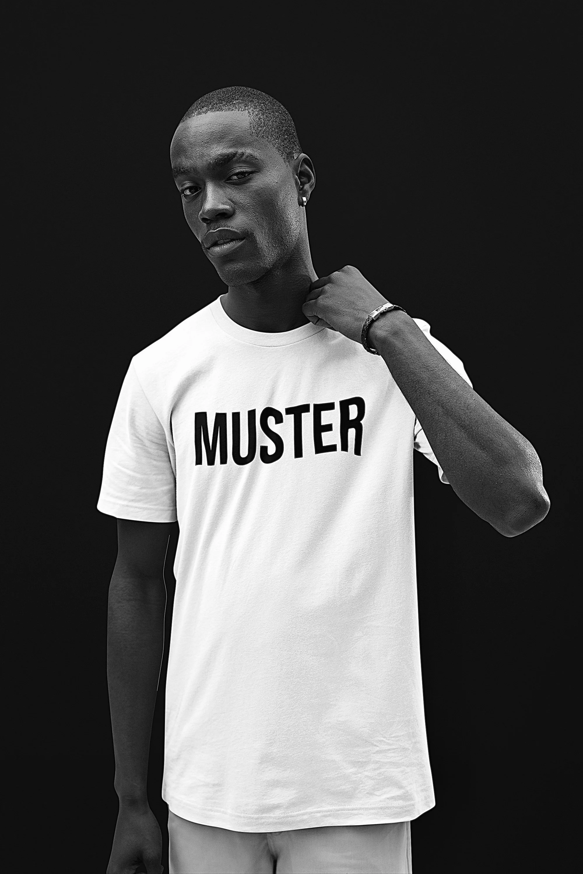 A young man with short hair and earrings is wearing a white T-shirt with the word 'MUSTER' printed on it. He is standing against a dark background and has one hand touching his neck.