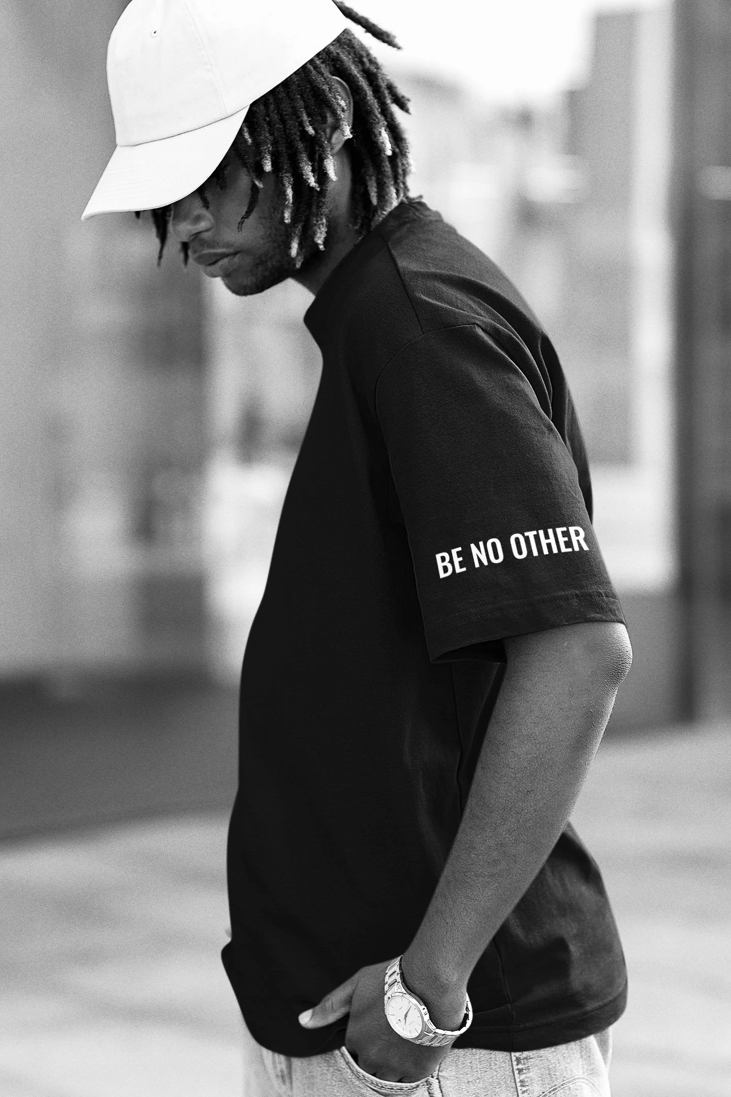 Black and white photo of a young man with dreadlocks wearing a baseball cap, a t-shirt with the phrase "BE NO OTHER" on the sleeve, and a wristwatch, standing outdoors with his head tilted downward.