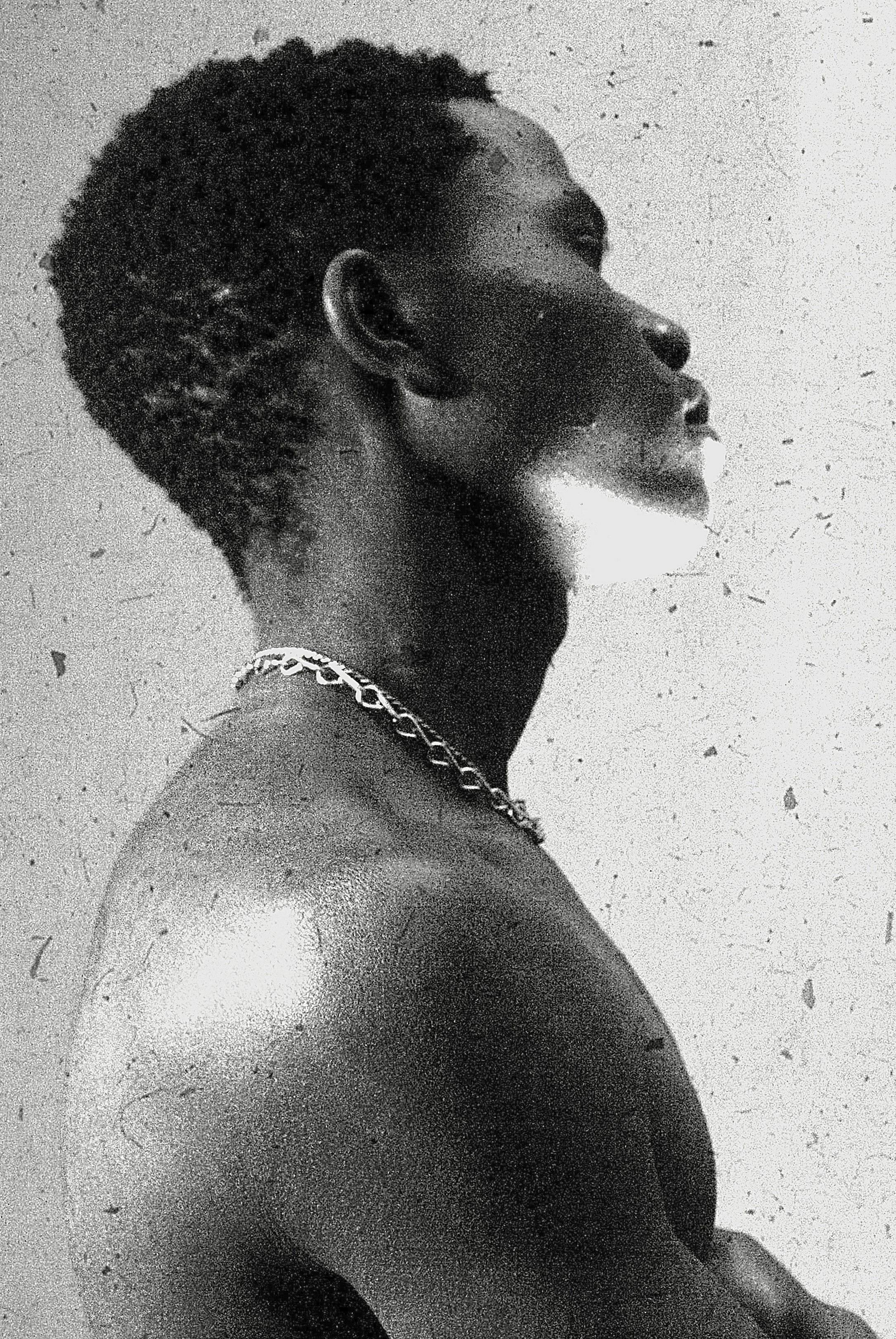 Black and white photograph of a Black man in profile, shirtless, wearing a chain necklace, with short textured hair, standing against a wall.