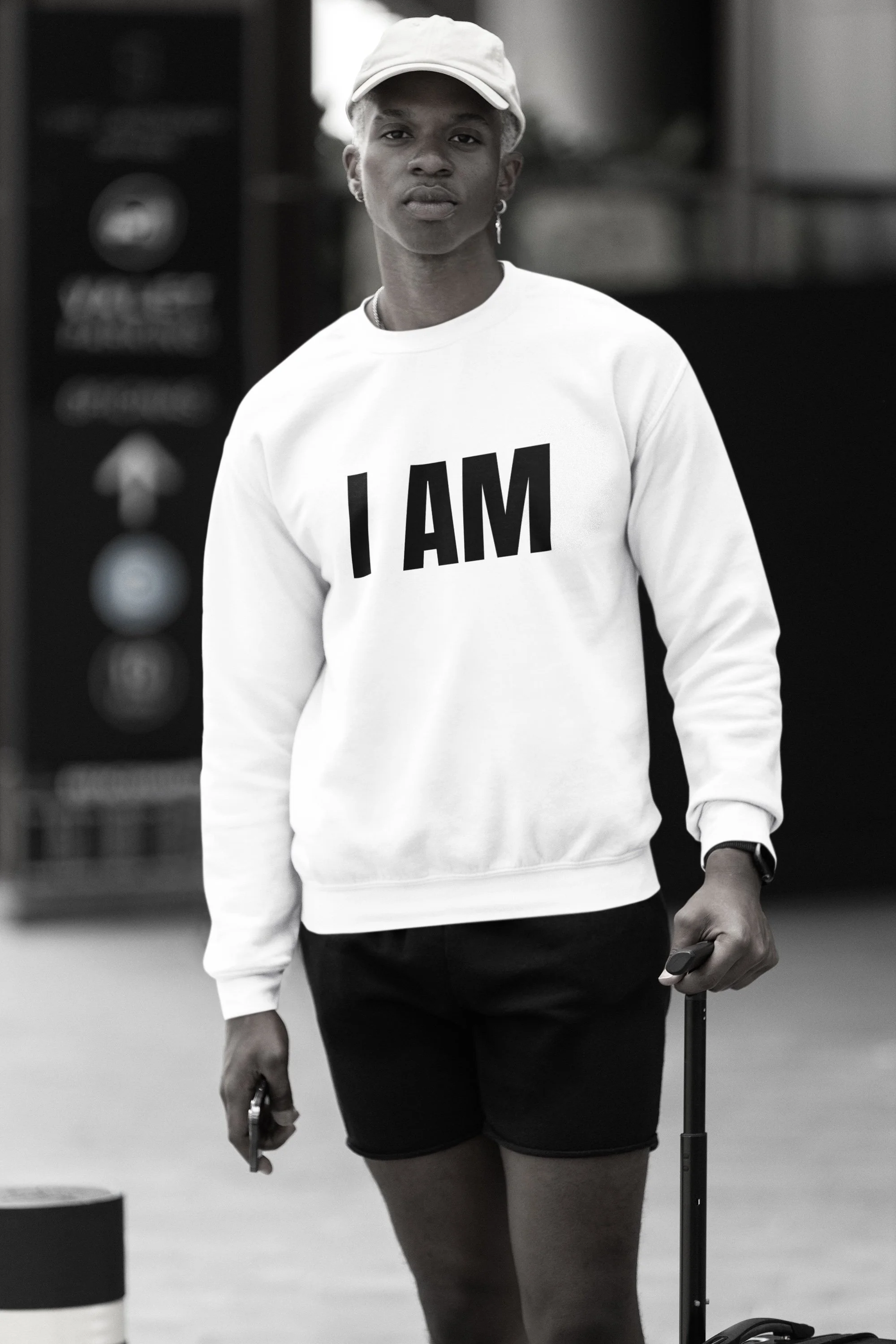 Black and white photo of a person with short curly hair, wearing a white cap, a white sweatshirt with 'I AM' written on it, black shorts, and holding a small suitcase and a phone.