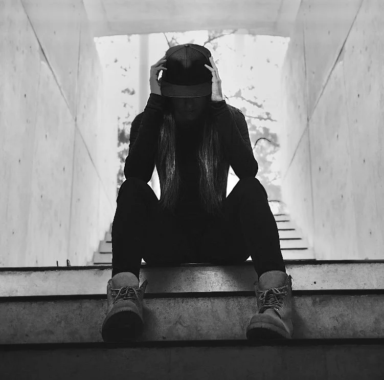 A person sitting on stairs, wearing a baseball cap and hoodie, holding the cap with both hands, with a dark silhouette against a bright background.