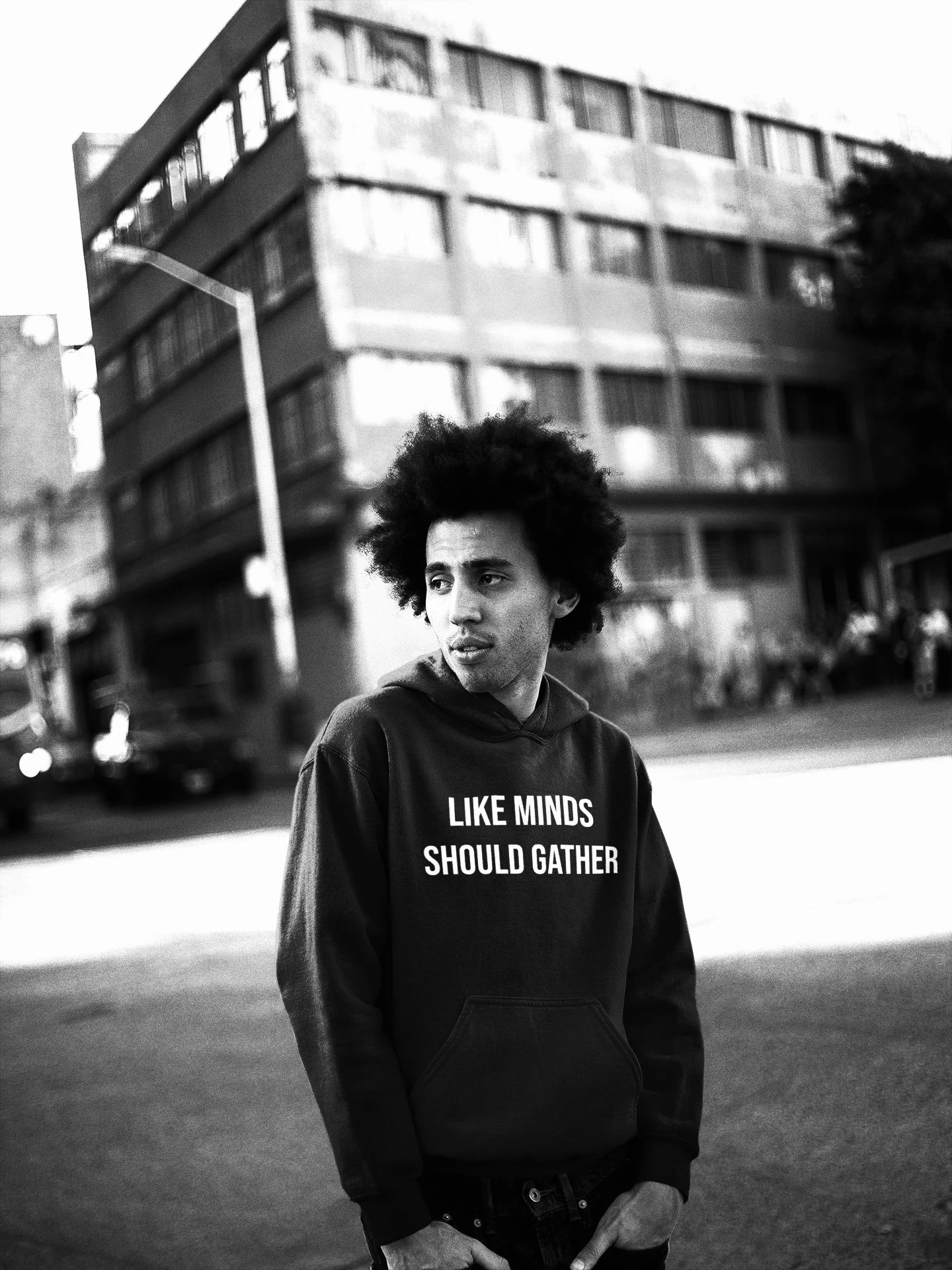 A young man with curly hair wearing a black hoodie with the phrase "LIKE MINDS SHOULD GATHER" standing outdoors on a city street near a large building, with cars and people in the background, in black and white.