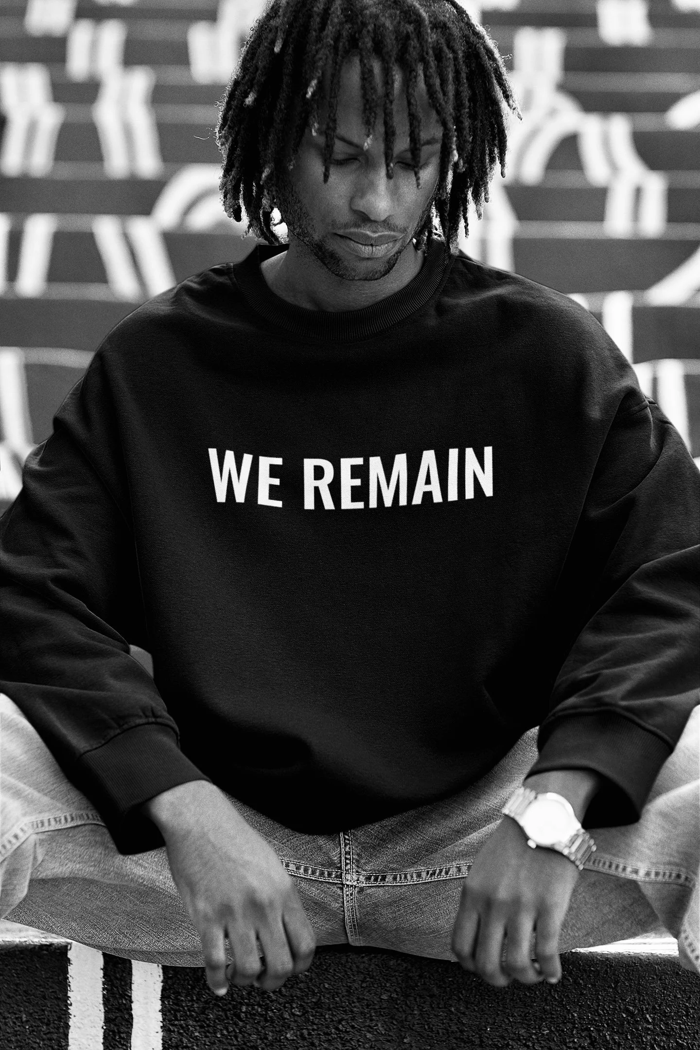 Black and white photo of a young man with dreadlocks, sitting cross-legged on a bench, wearing a black sweatshirt with the words 'WE REMAIN' on it, and looking down.