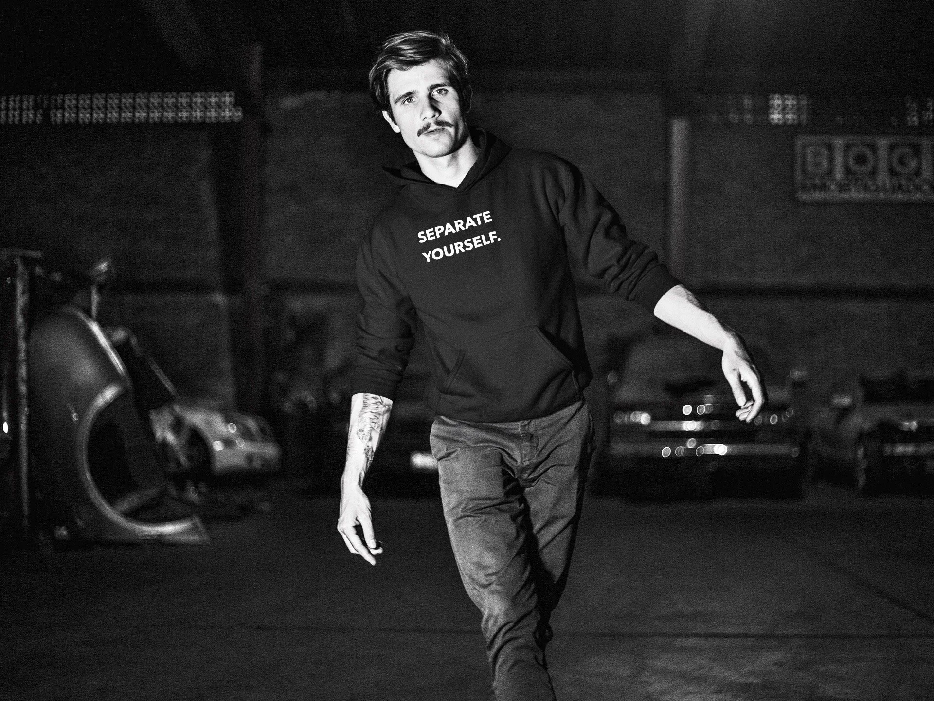 A young man with a mustache and tattoos on his arms is walking in a parking garage at night. He is wearing a black hoodie with the words "SEPARATE YOURSELF" printed on it and dark jeans.