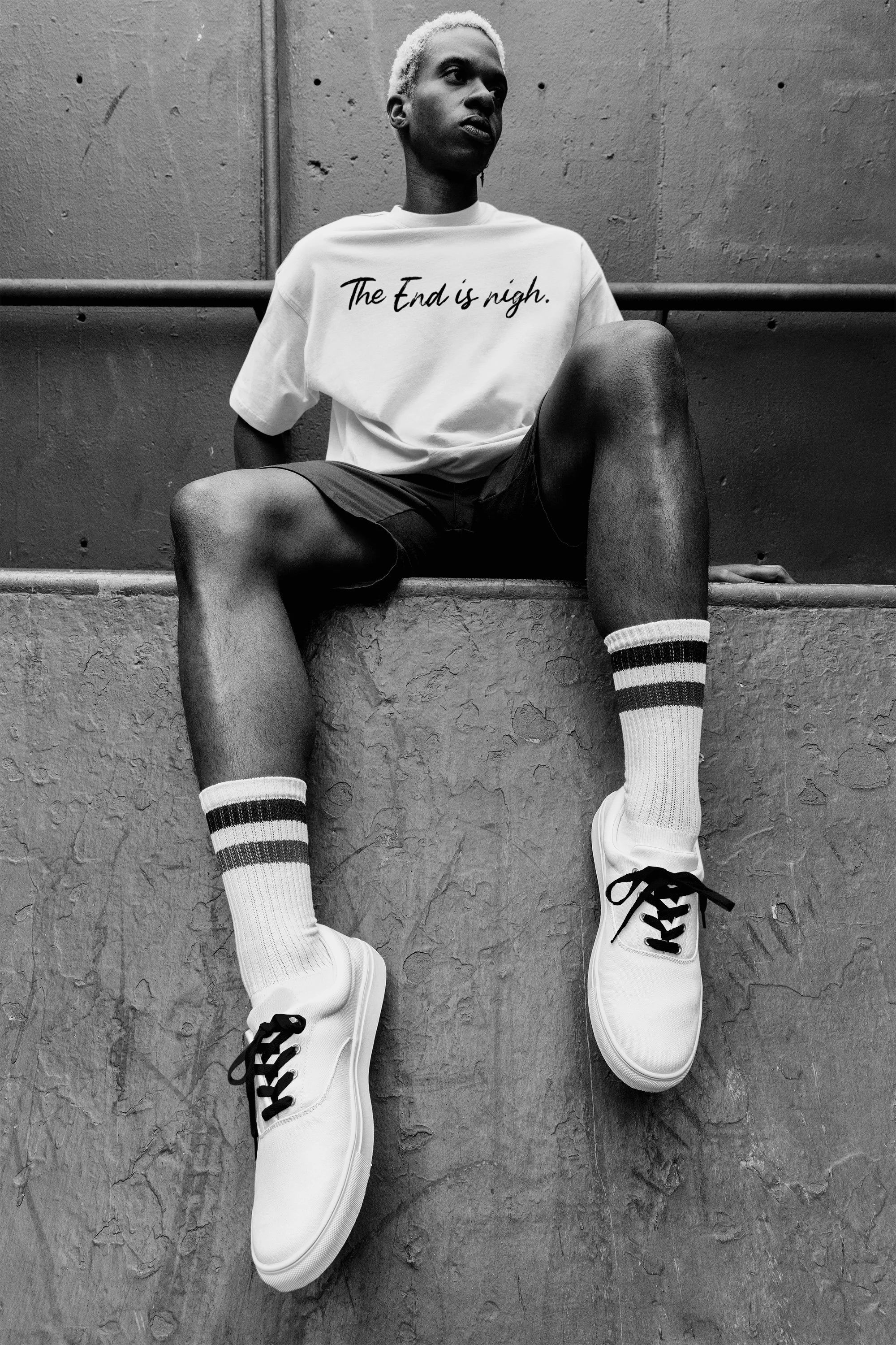 A person with short blonde hair sitting on a ledge, wearing a t-shirt that says "The End is nigh", shorts, striped knee-high socks, and white sneakers, against a concrete wall.