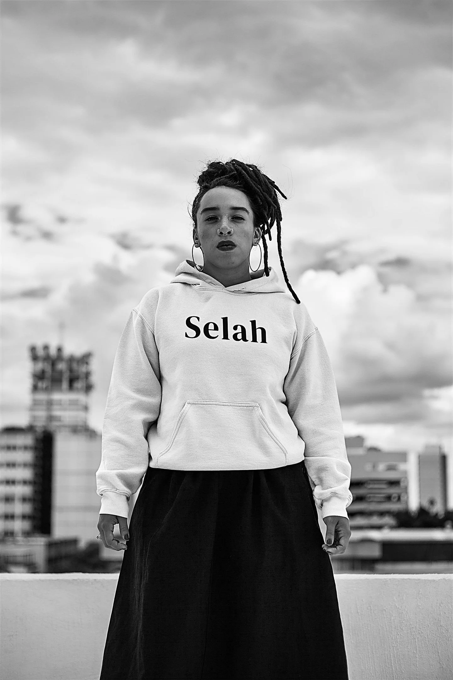 Black and white photo of a woman with dreadlocks wearing a hoodie with the name "Selah" on it, standing outdoors with a cityscape and cloudy sky in the background.