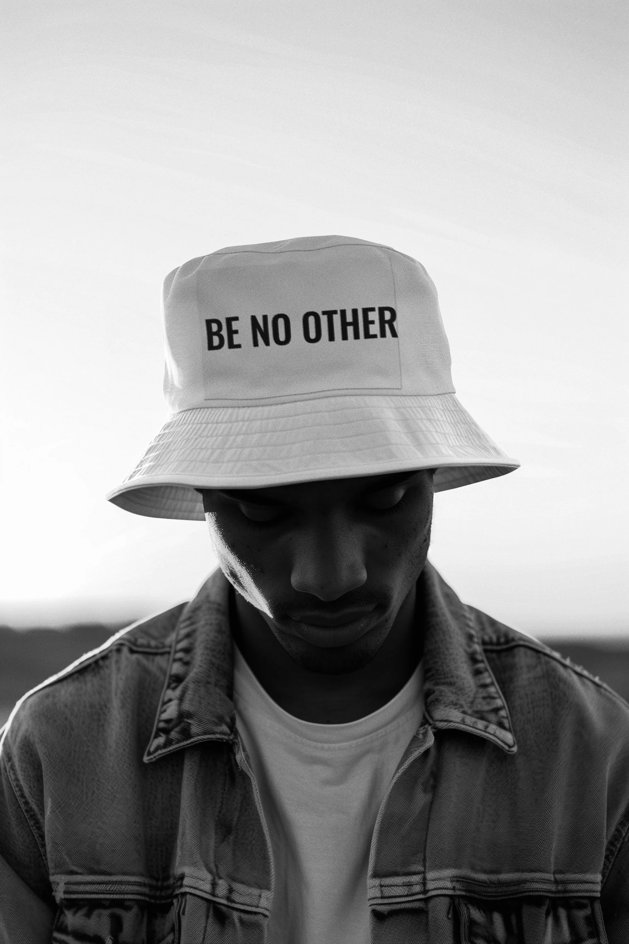 A close-up black and white photo of a young man wearing a bucket hat with the words "BE NO OTHER" printed on it. He is looking downward, wearing a denim jacket and a white t-shirt, with a blurred outdoor background.