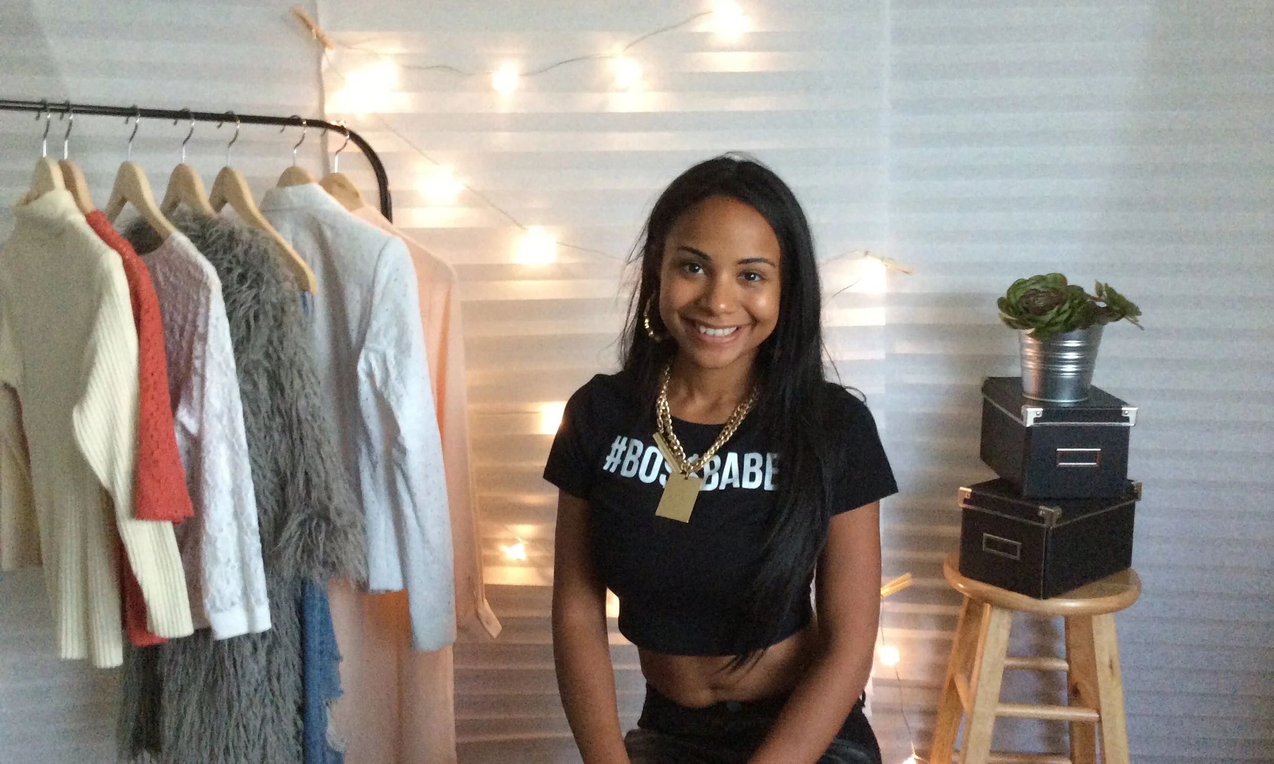 A young woman with long black hair smiling and wearing a black t-shirt with '#BOSSBABE' written on it, sitting in a room with a clothing rack, string lights, a wooden stool, and a potted plant in the background.