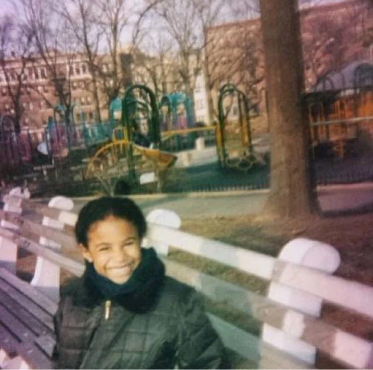 A smiling child sitting on a park bench with a playground and trees in the background.
