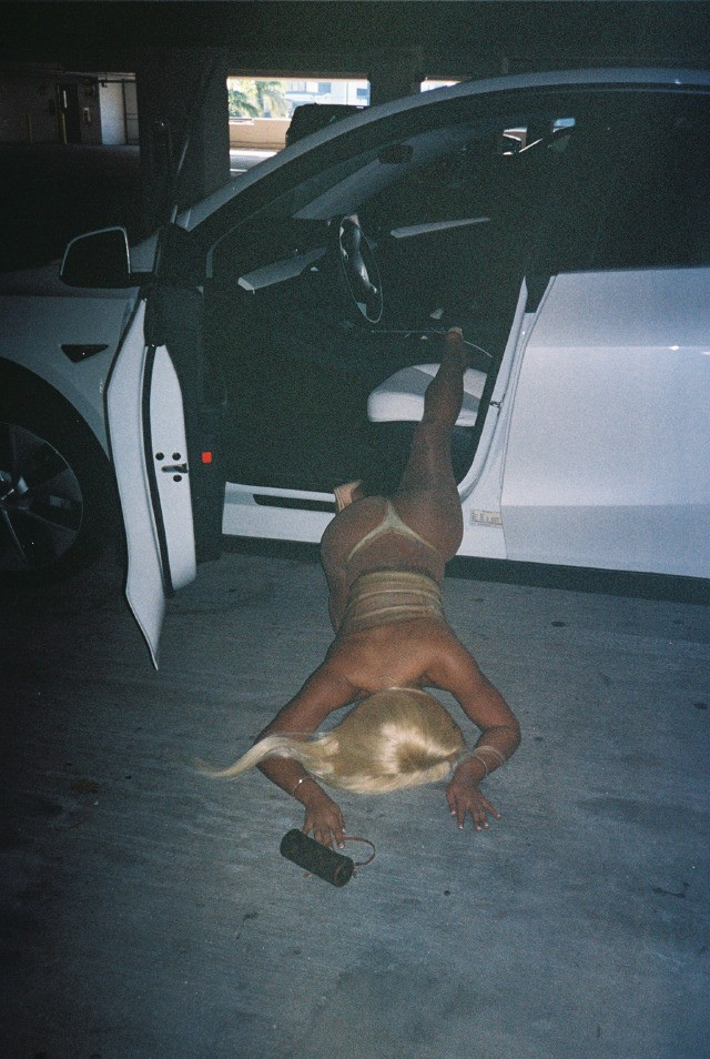 Person doing a headstand with feet on the car door sill in a parking garage, holding an object in one hand.