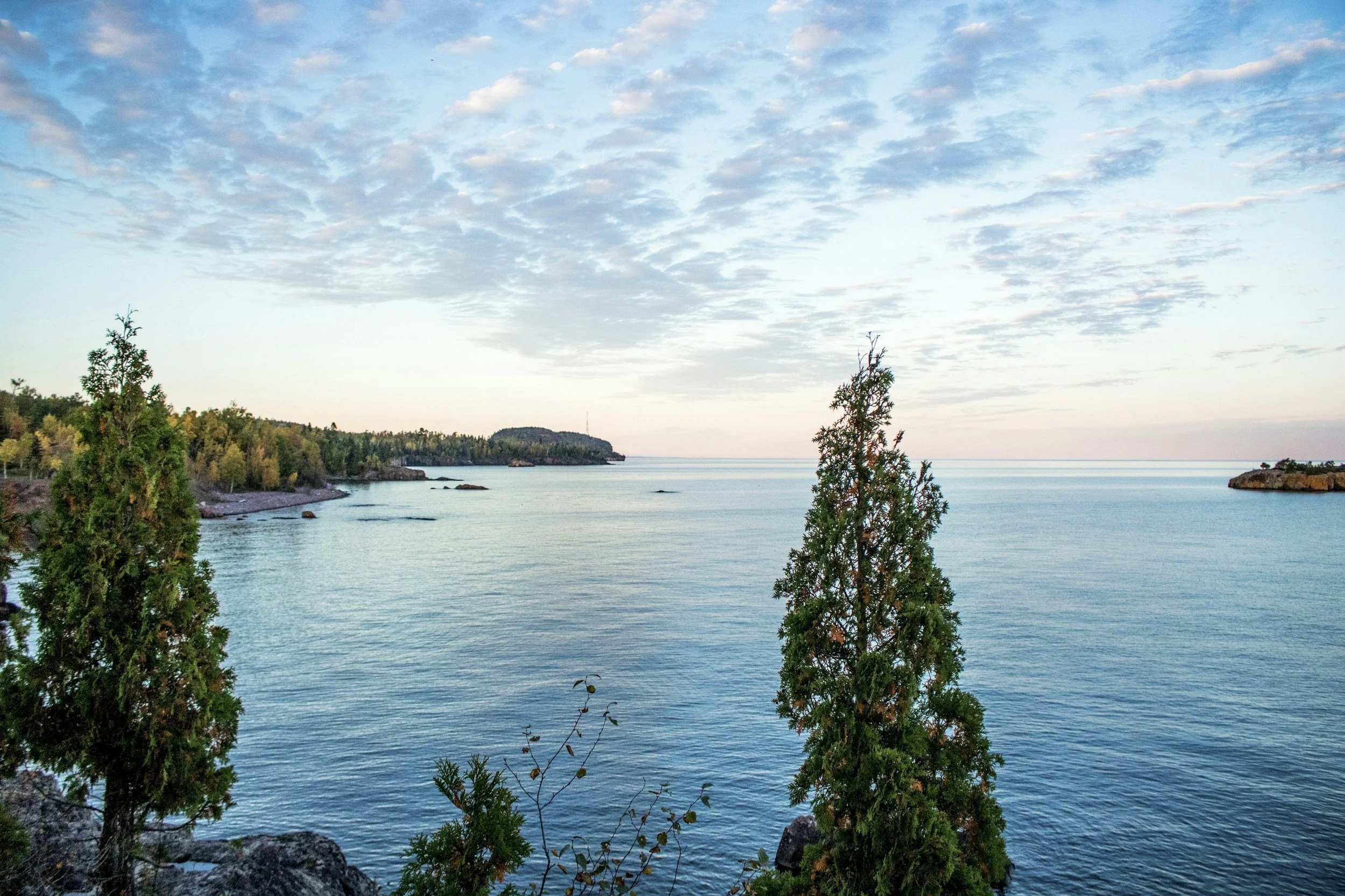 A calm body of water, possibly a bay or lake, surrounded by trees and rocky shores under a sky with scattered clouds.