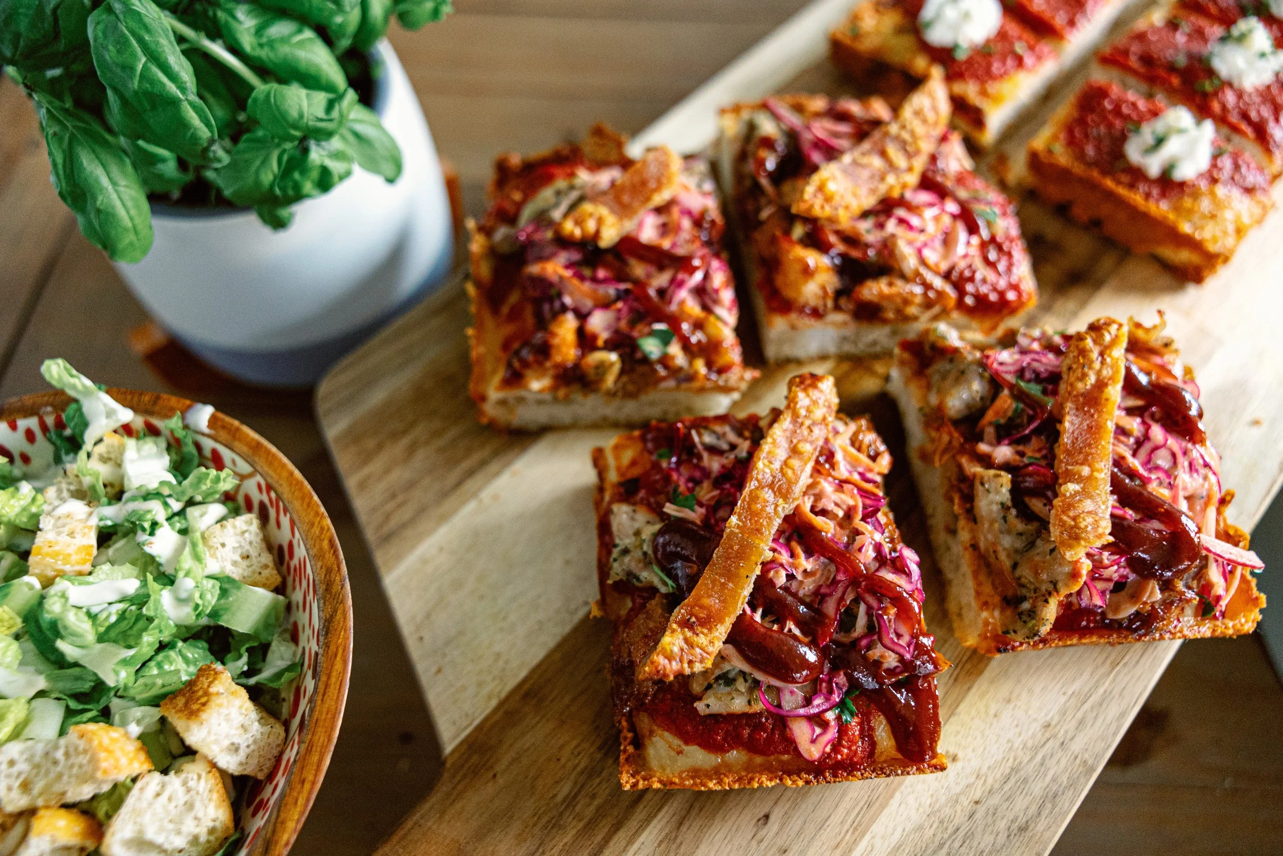 Slices of Detroit pizza with pulled pork and coleslaw on a wooden serving board, beside a bowl of Caesar salad.