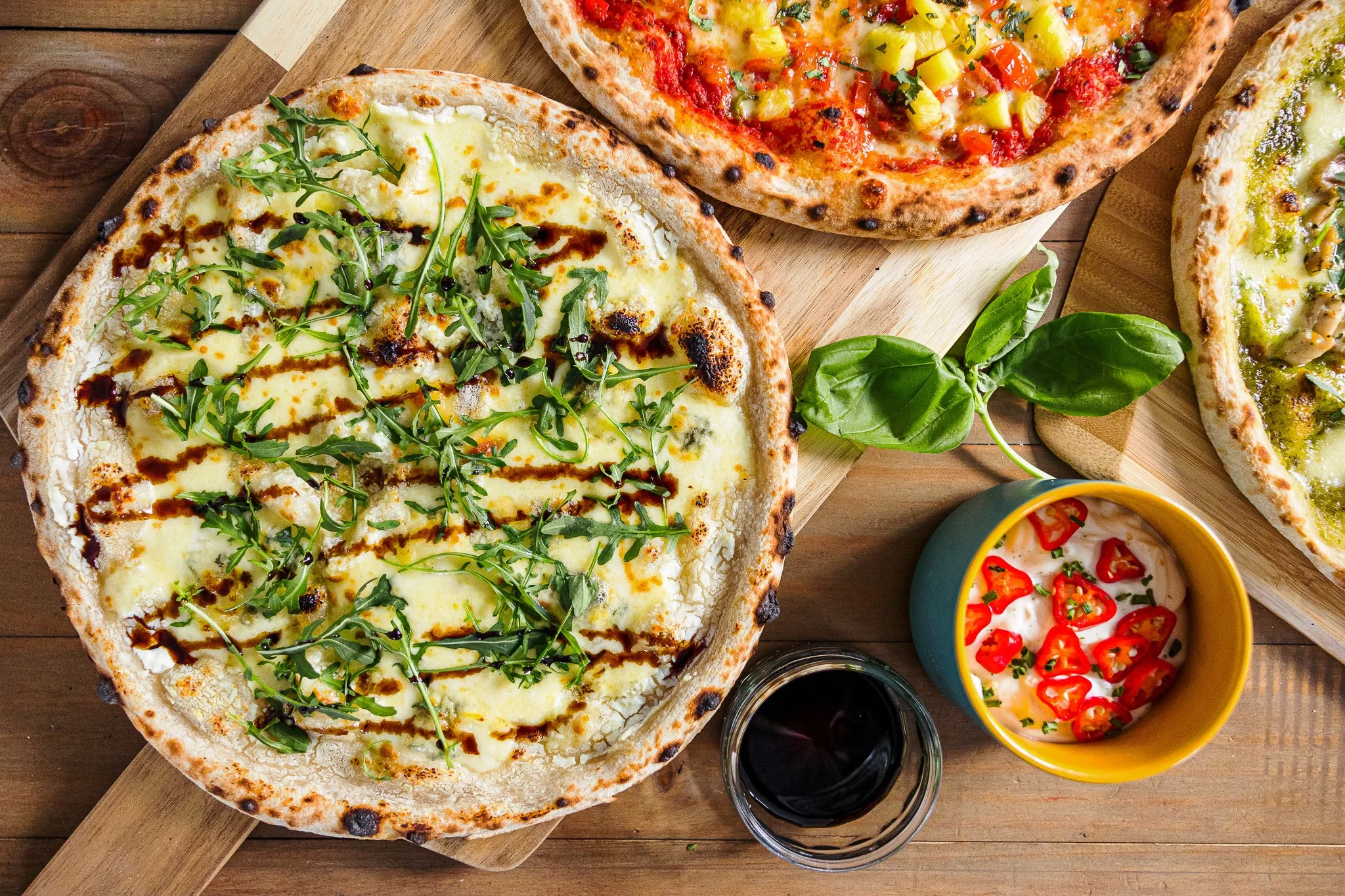 Three different types of pizzas on wooden boards with a small bowl of sauce, a glass of red wine, and a bowl of creamy dip topped with red chili slices, basil, and herbs, set on a wooden table.