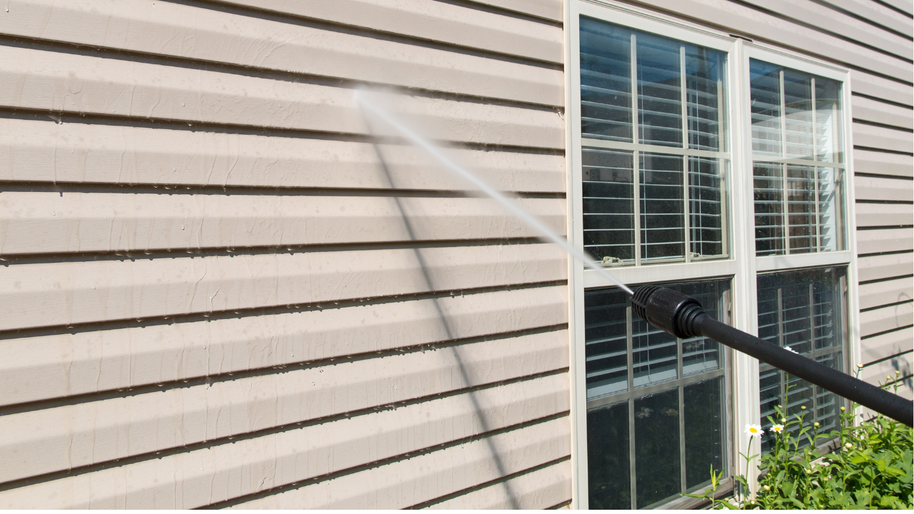 A person using a high-pressure water hose to clean the exterior wall of a house with white siding and a multi-pane window.