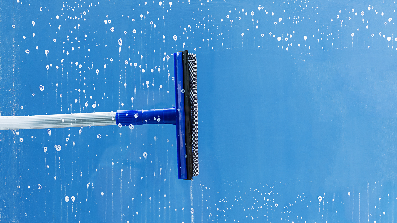 A blue squeegee cleaning a glass window with soap suds and water streaks.
