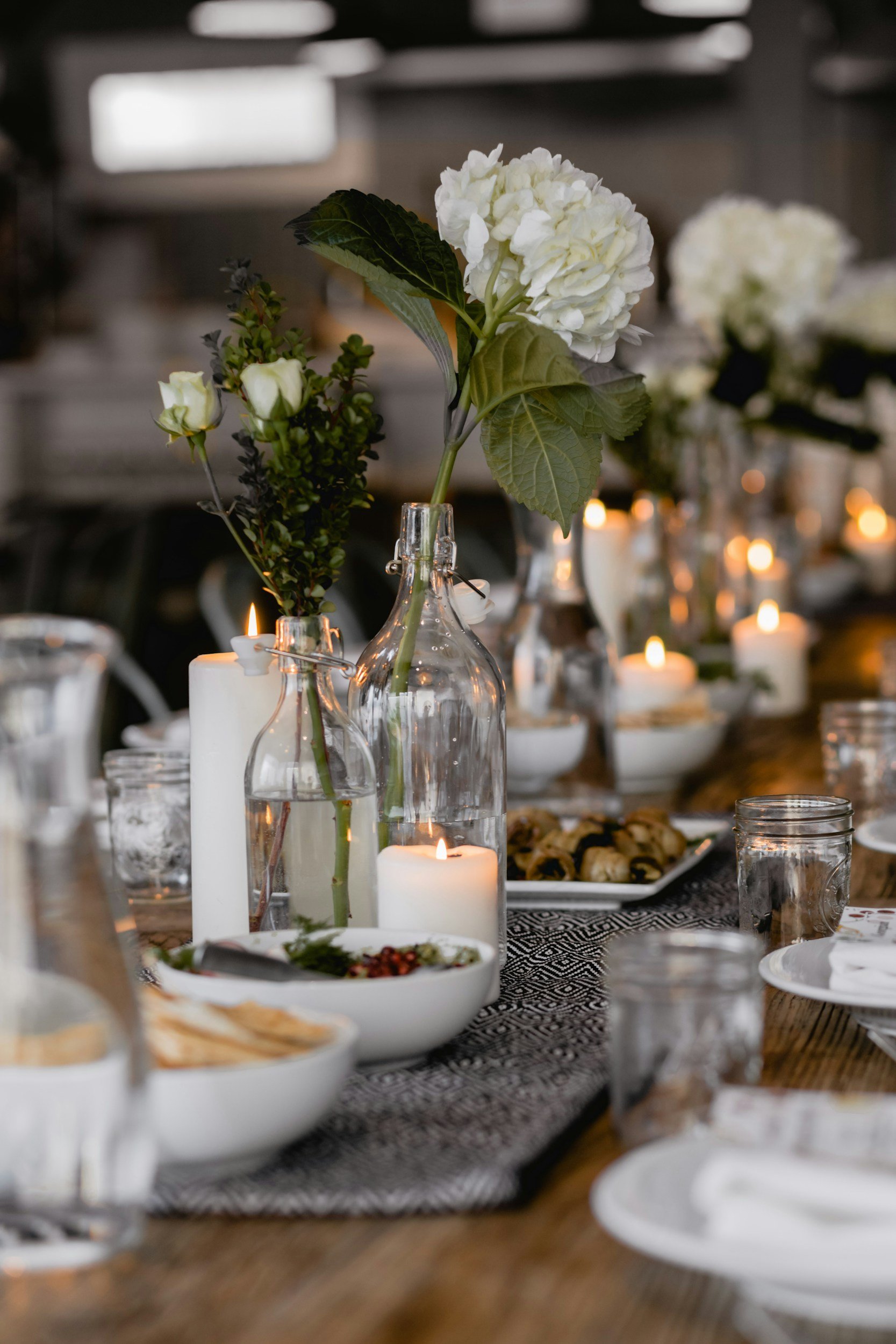 Elegant dining table with white flowers in glass bottles, lit candles, plates of food, and drinking glasses, set against a dimly lit background.