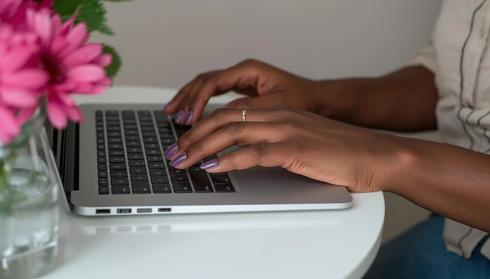 Hands typing on a laptop keyboard on a white table, with a pink flower in a glass vase nearby.
