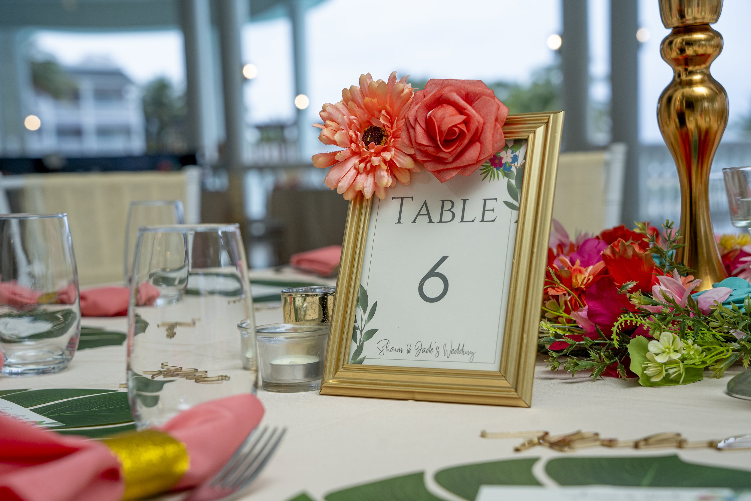Table number 6 at a wedding reception with pink and green floral decorations, pink napkins, water glasses, and gold accents, in a room with large windows and natural light.