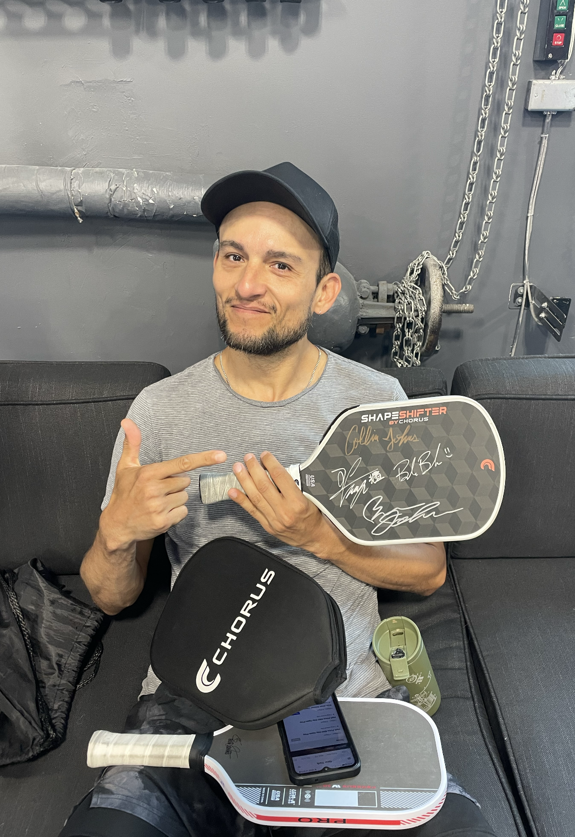 A man sitting on a black couch in a sports facility, holding a pickleball paddle with signatures, pointing at it, with a black CHORUS bag, a smartphone, a water bottle, and a hockey stick on his lap.
