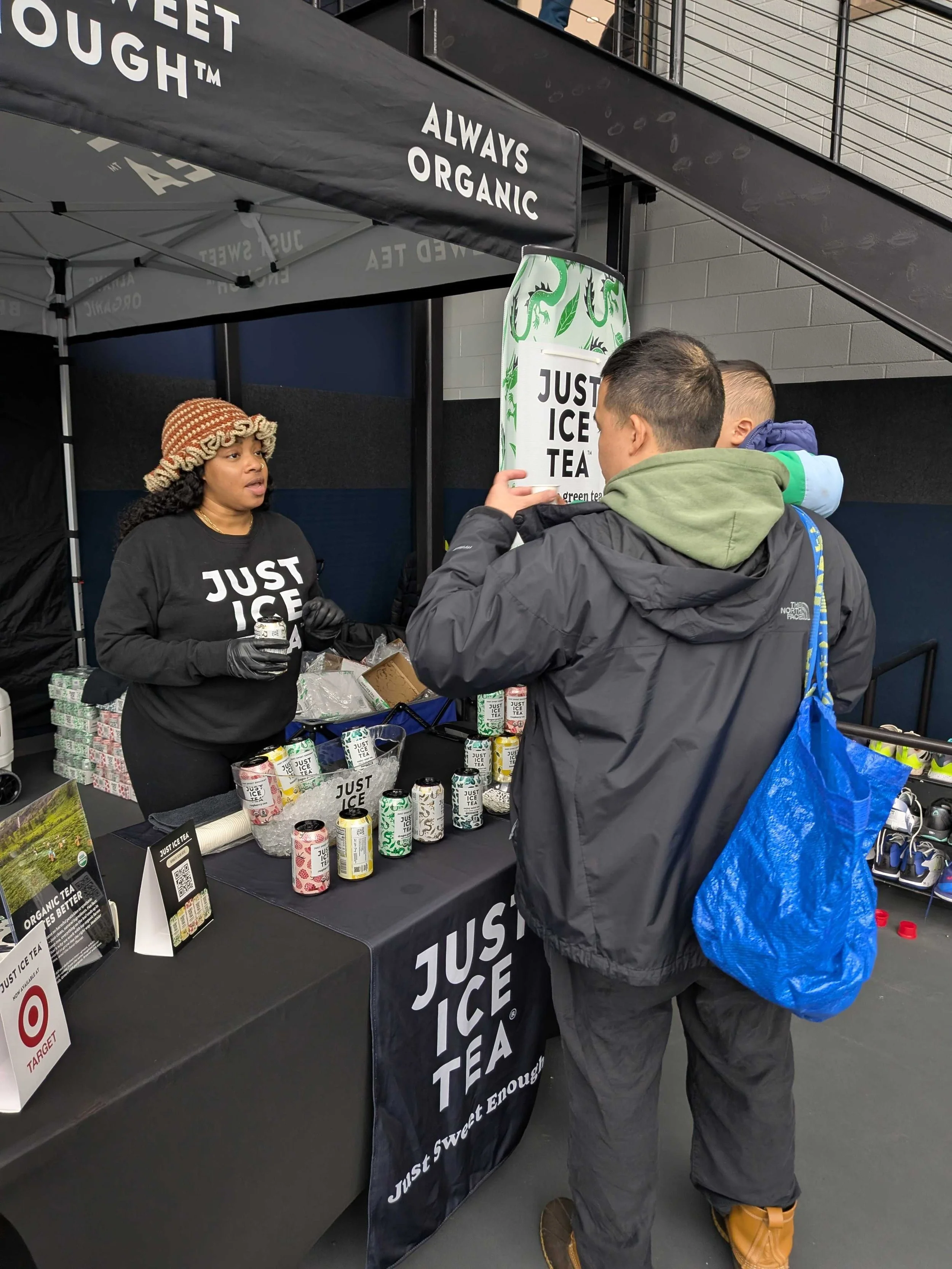 A woman at a booth selling Just Ice Tea. She is wearing a black shirt with white text, a brown and white knit hat, and black gloves. There are cans of different flavors of Just Ice Tea on the table. Two men are buying tea; one is wearing a green hoodie and a black jacket, holding a blue bag, while the other wears a blue jacket. The booth is shaded by a canopy with the words 'Always Organic' and 'Sweet Enough.'