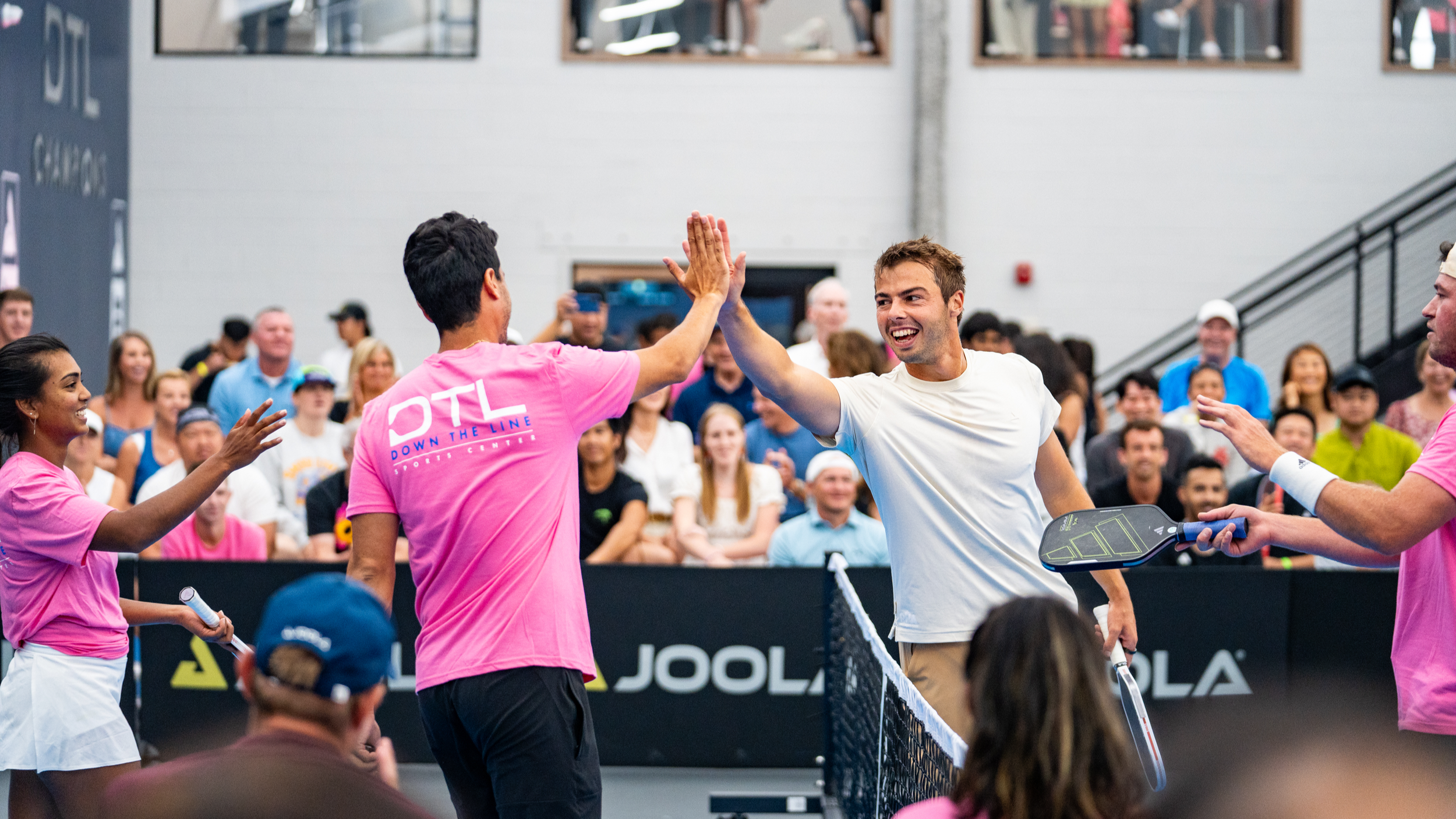 Two tennis players in pink shirts high-fiving at the net after a match, with a smiling male player in a white shirt holding a tennis racket, and a crowd cheering in the background at an indoor tennis stadium.