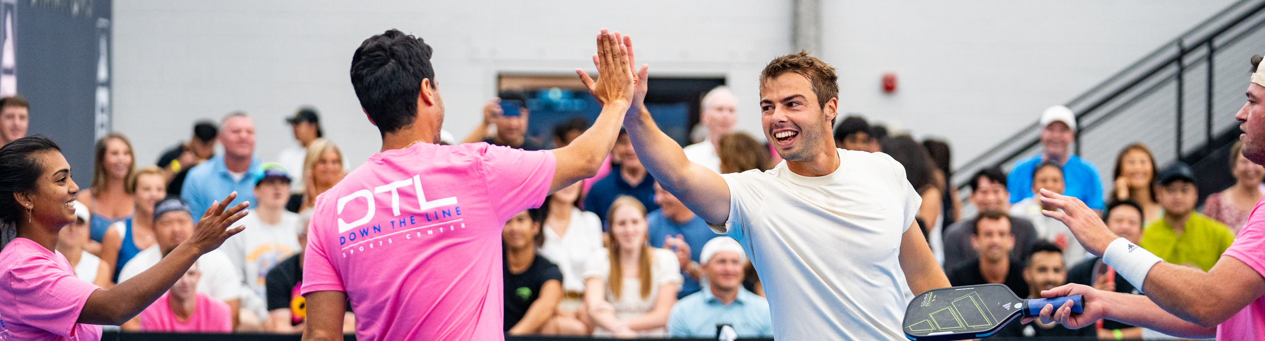 Two men high-five after a tennis match indoors, with a crowd watching and smiling in the background.