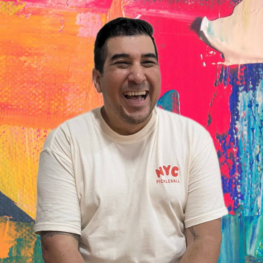 A smiling man with dark hair and a light-colored T-shirt with NYC Pickleball logo, sitting in front of a colorful abstract painting background.