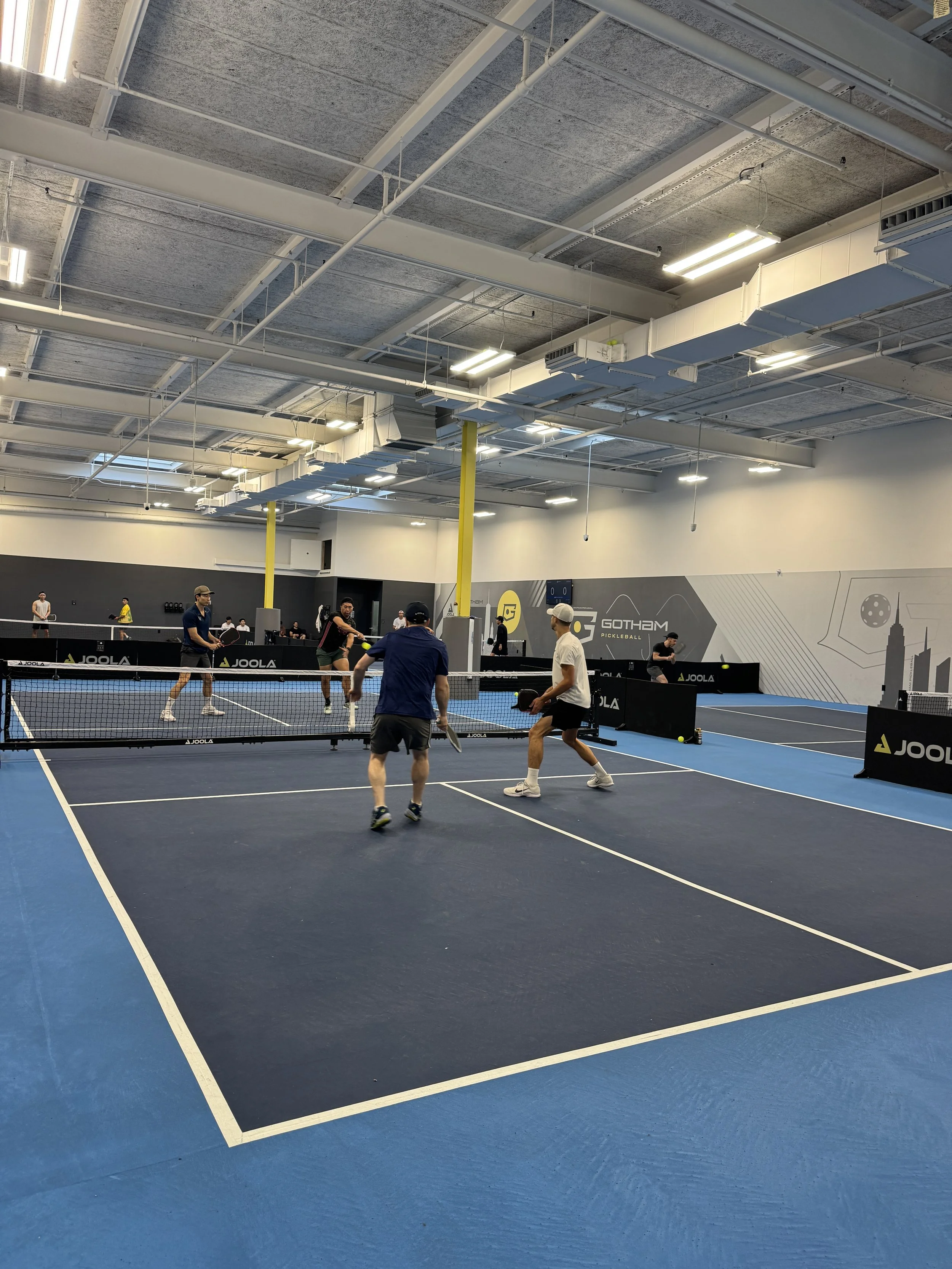 People playing pickleball in an indoor sports facility with multiple courts, blue flooring, and high ceiling.