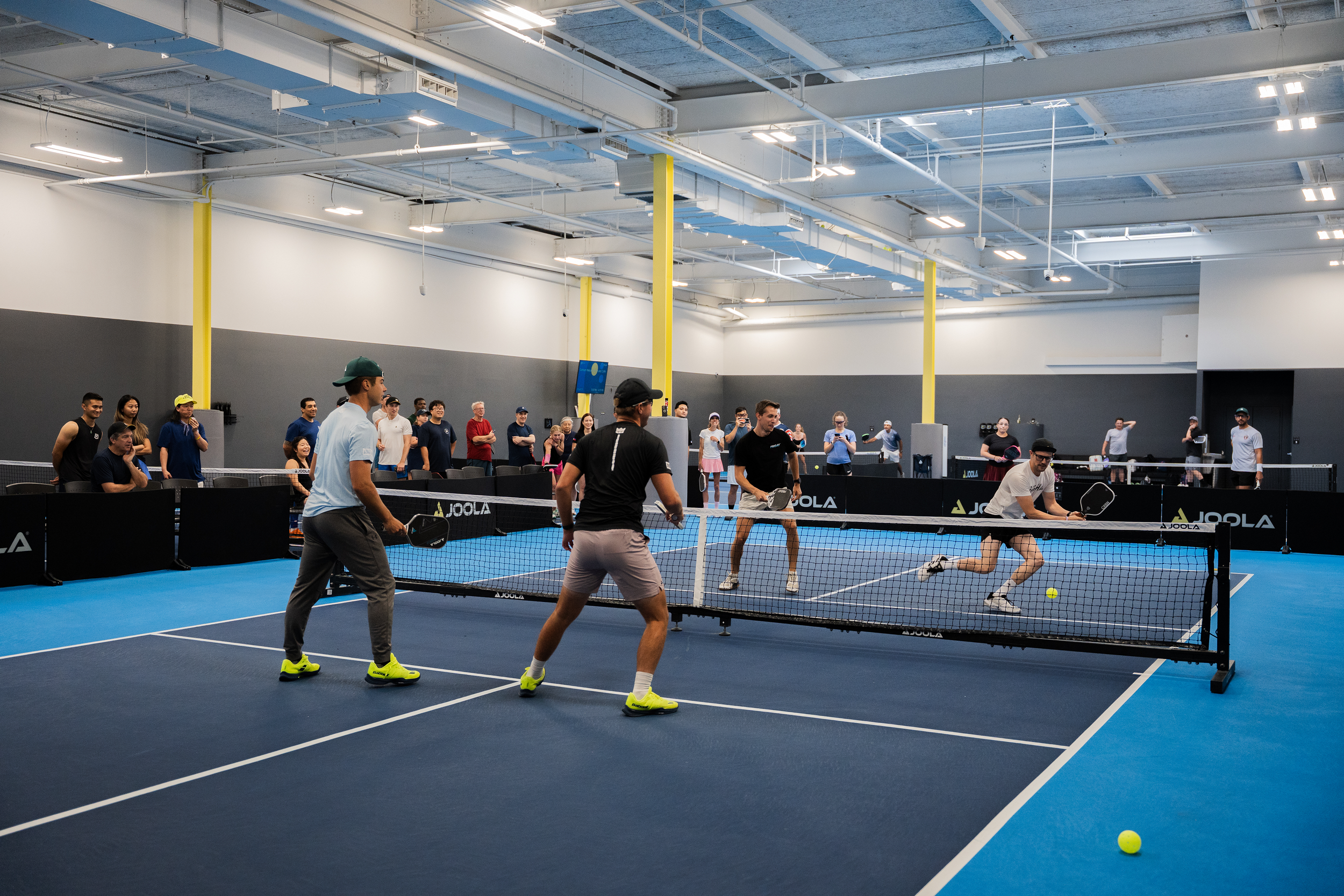 People playing pickleball on indoor court surrounded by spectators.