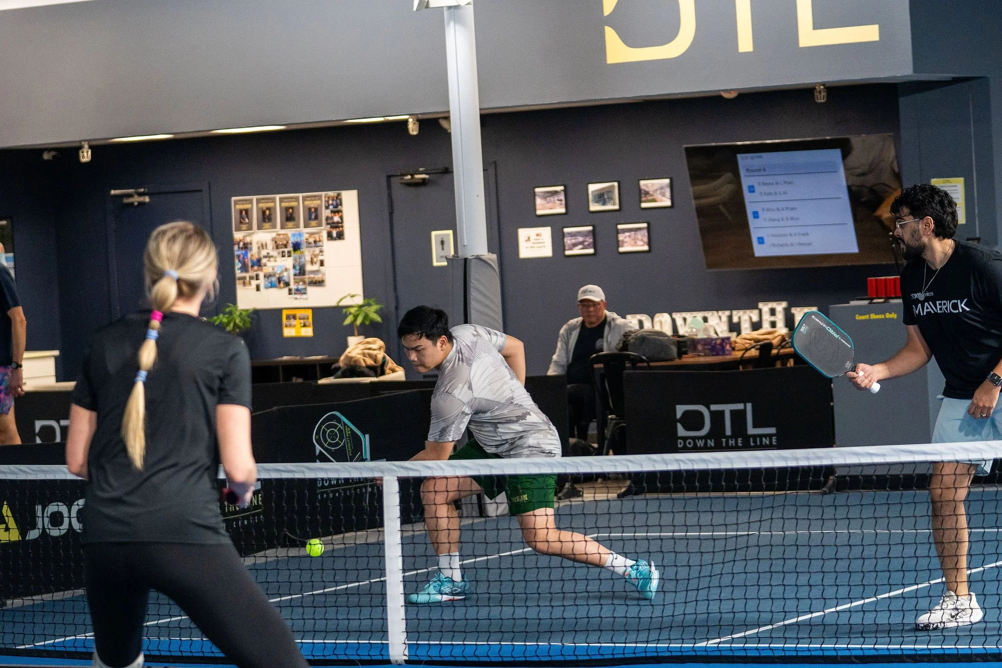 People playing pickleball indoors, with a woman preparing to hit the ball and a man holding a paddle on the court.
