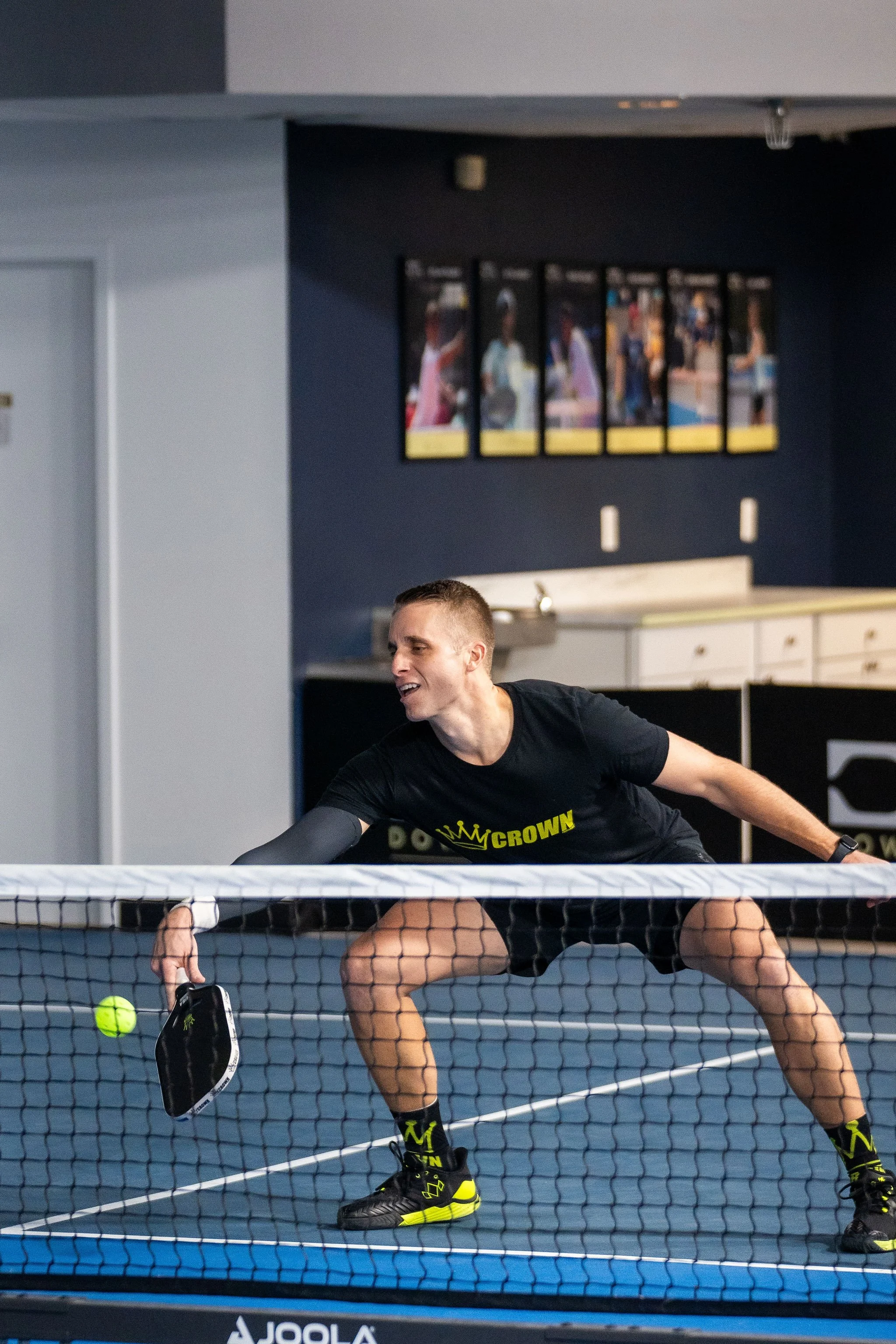 A man playing pickleball indoors, holding a paddle and hitting a neon green ball over the net.