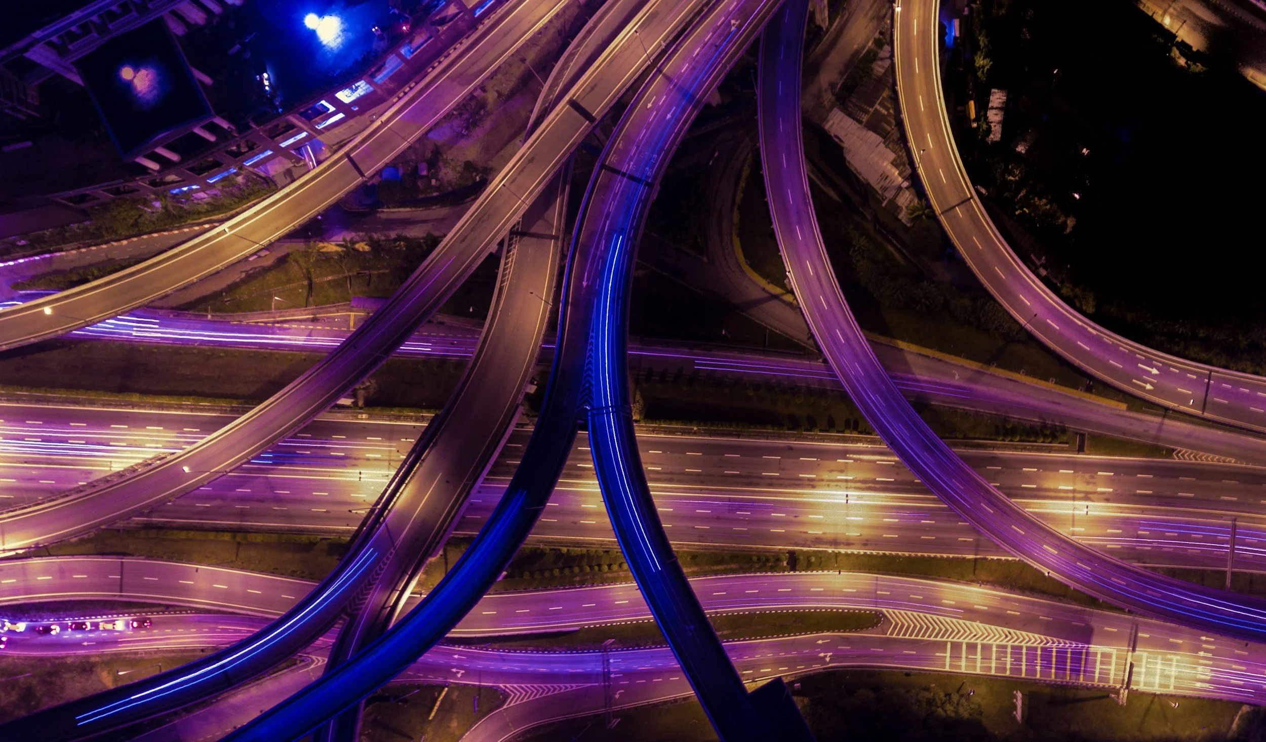 Night aerial view of a highway interchange with colorful purple and blue lighting illuminating the roads.