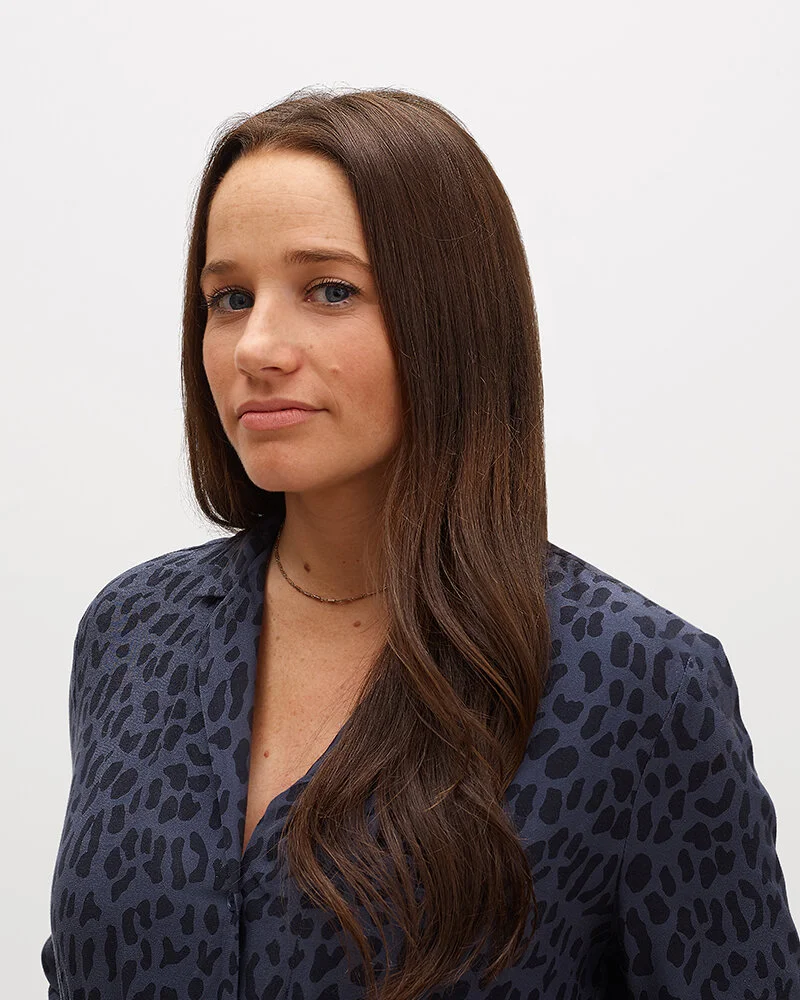 A woman with long brown hair wearing a dark animal print blouse against a plain white background.