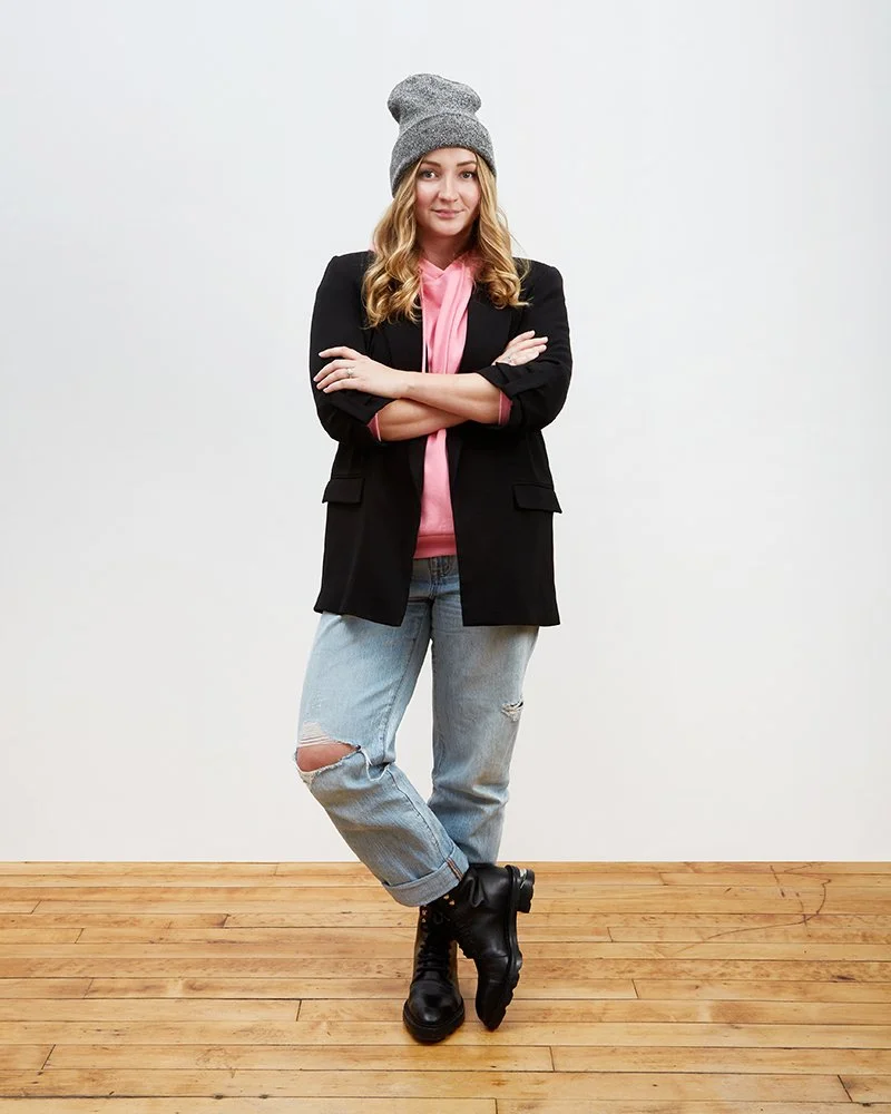 A young woman standing against a white background with crossed arms, wearing a gray beanie, pink hoodie, black blazer, ripped jeans, and black combat boots.