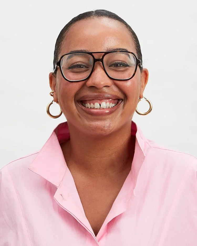 A woman with short hair wearing glasses, gold hoop earrings, and a pink collared shirt, smiling against a plain white background.